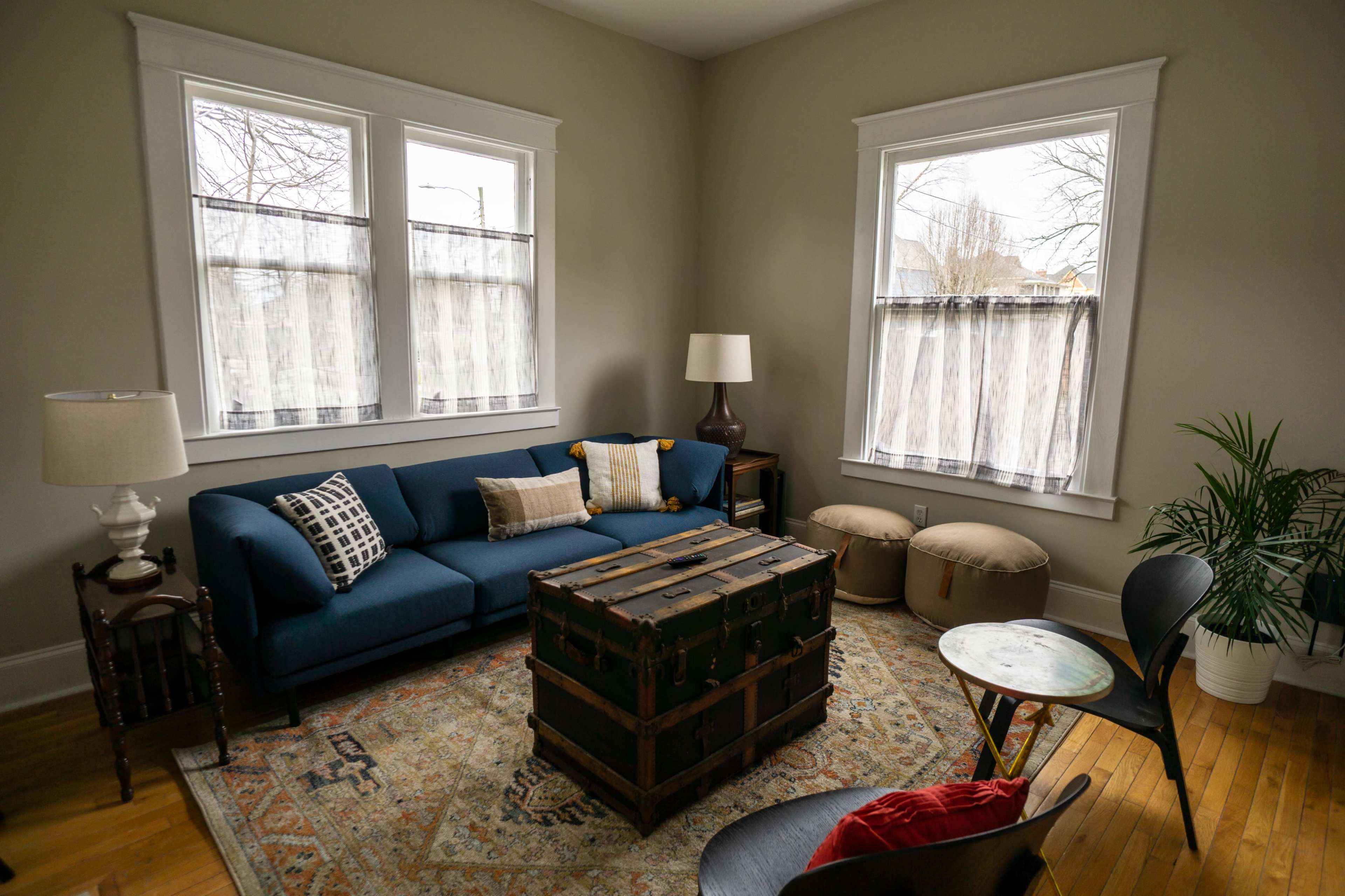 The image shows a living room featuring a blue couch, a wooden trunk as a coffee table, and two poufs, with natural light coming through the windows.
