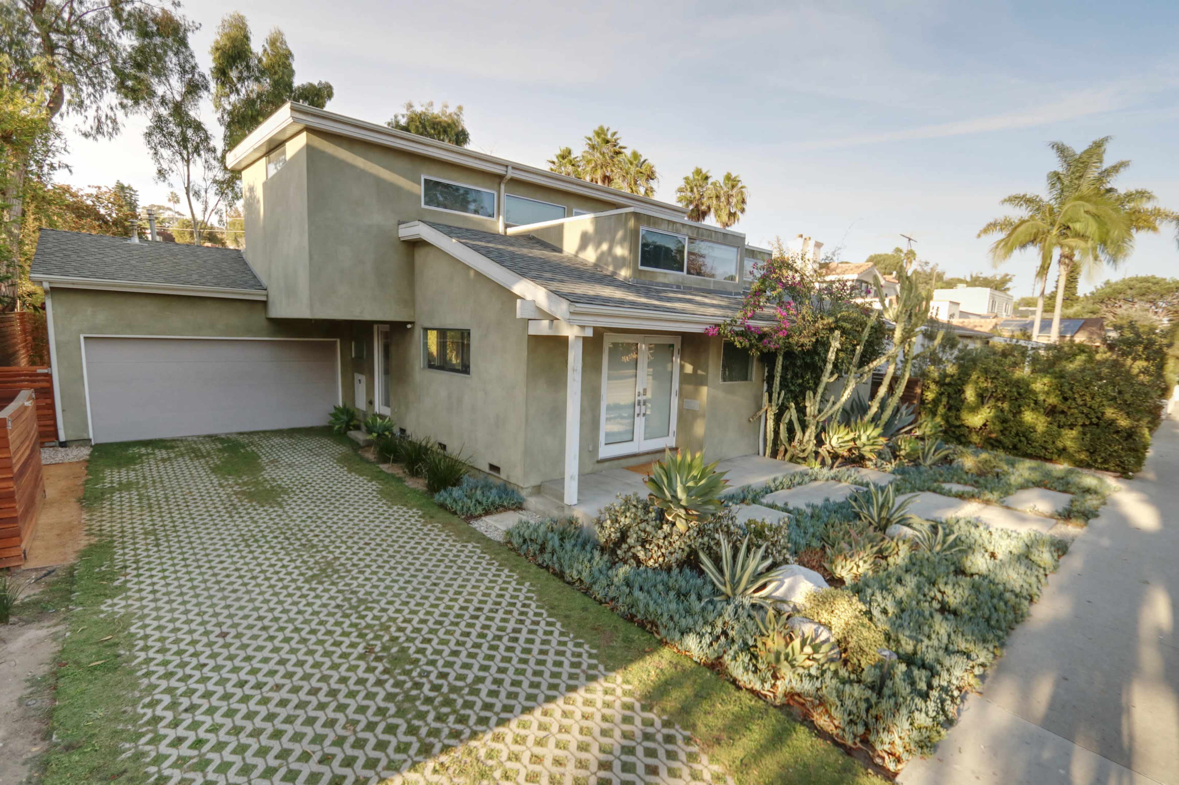 A modern two-story house with a landscaped front yard featuring various plants and a unique driveway pattern.