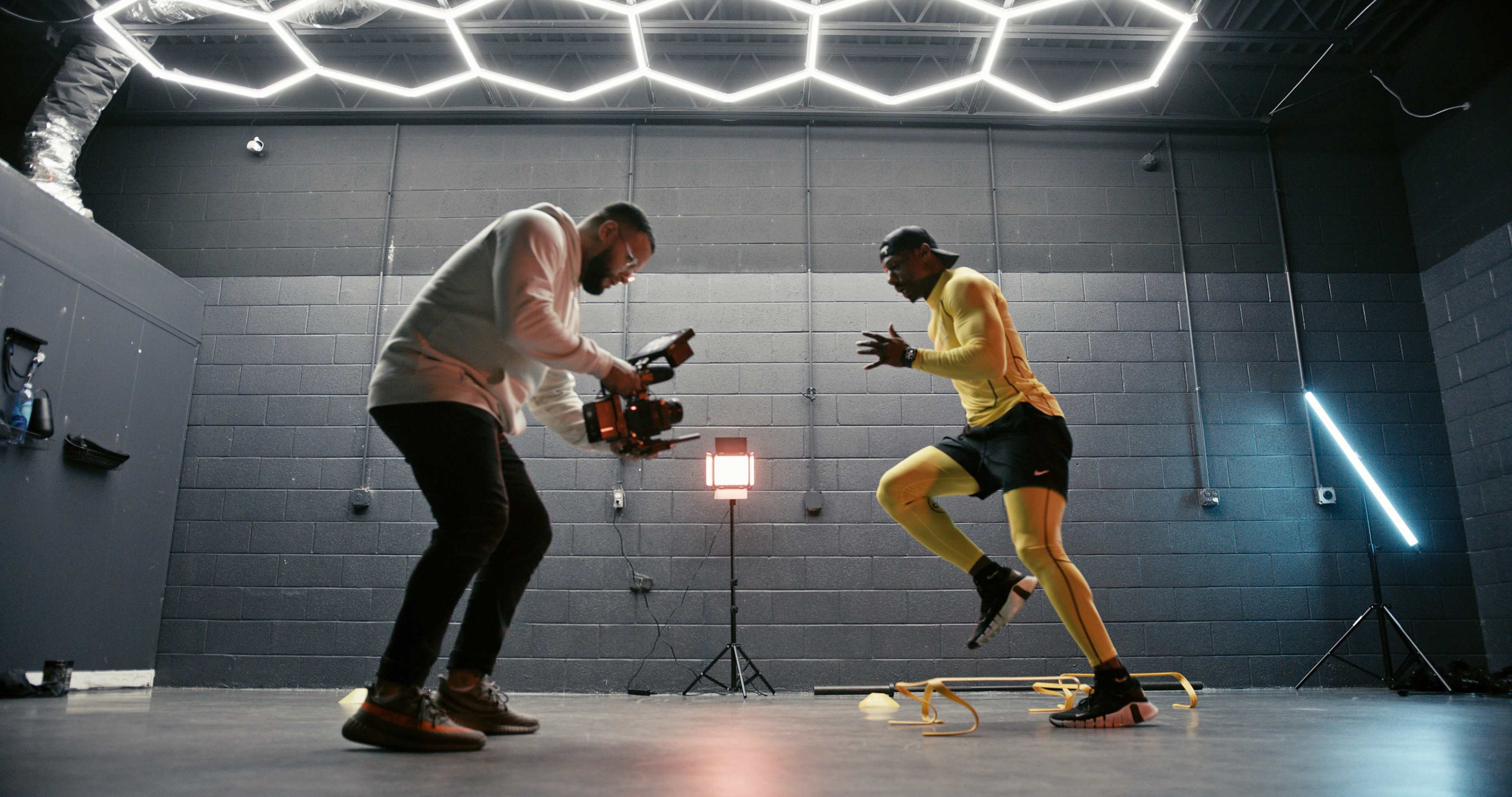A man in a yellow athletic outfit performs agility drills while another man films him with a camera in a visually striking indoor gym.