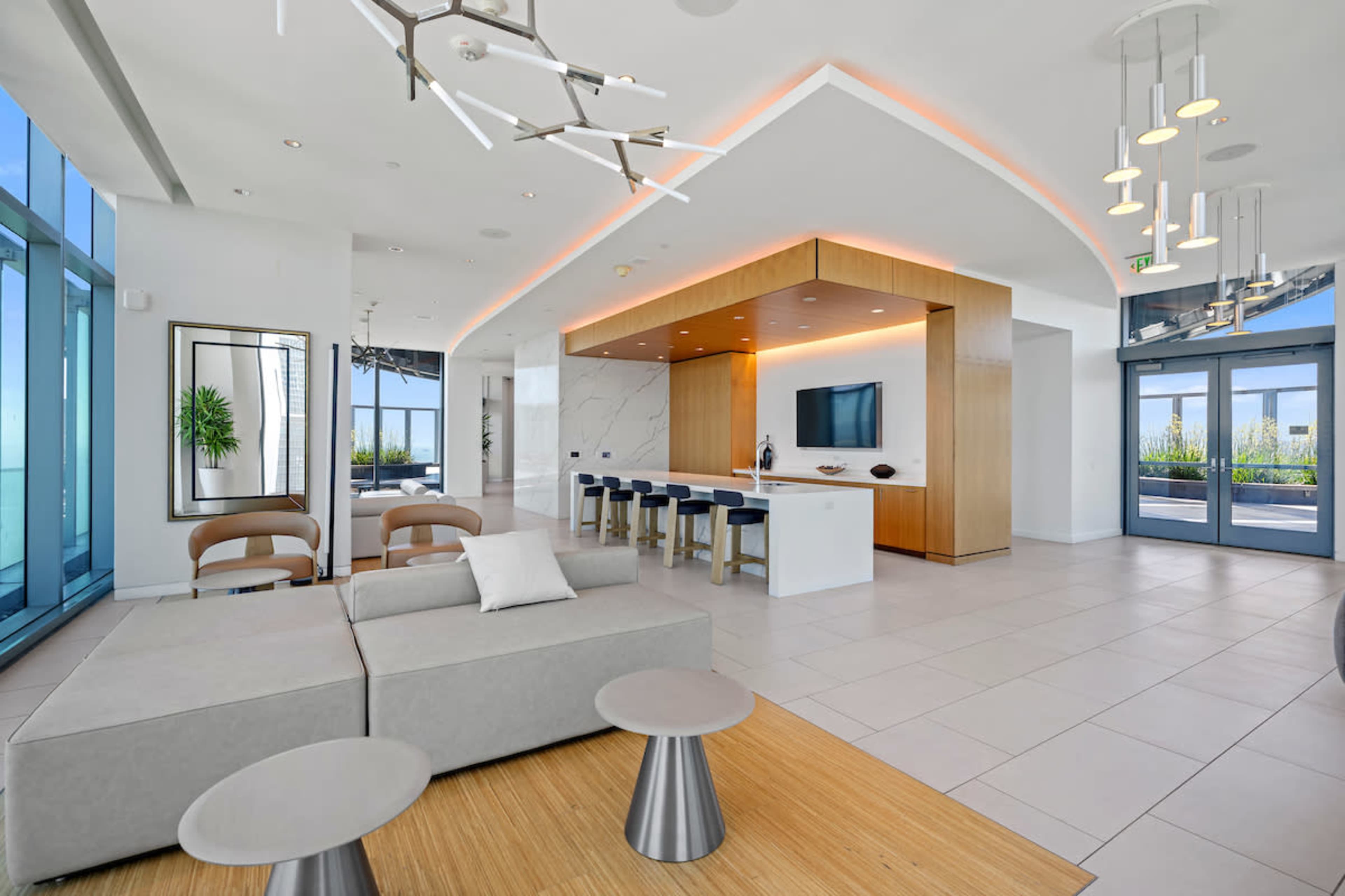 The image shows a modern, open-concept lounge area featuring a light-colored sofa, a bar counter with stools, and large windows offering views of the outdoors.