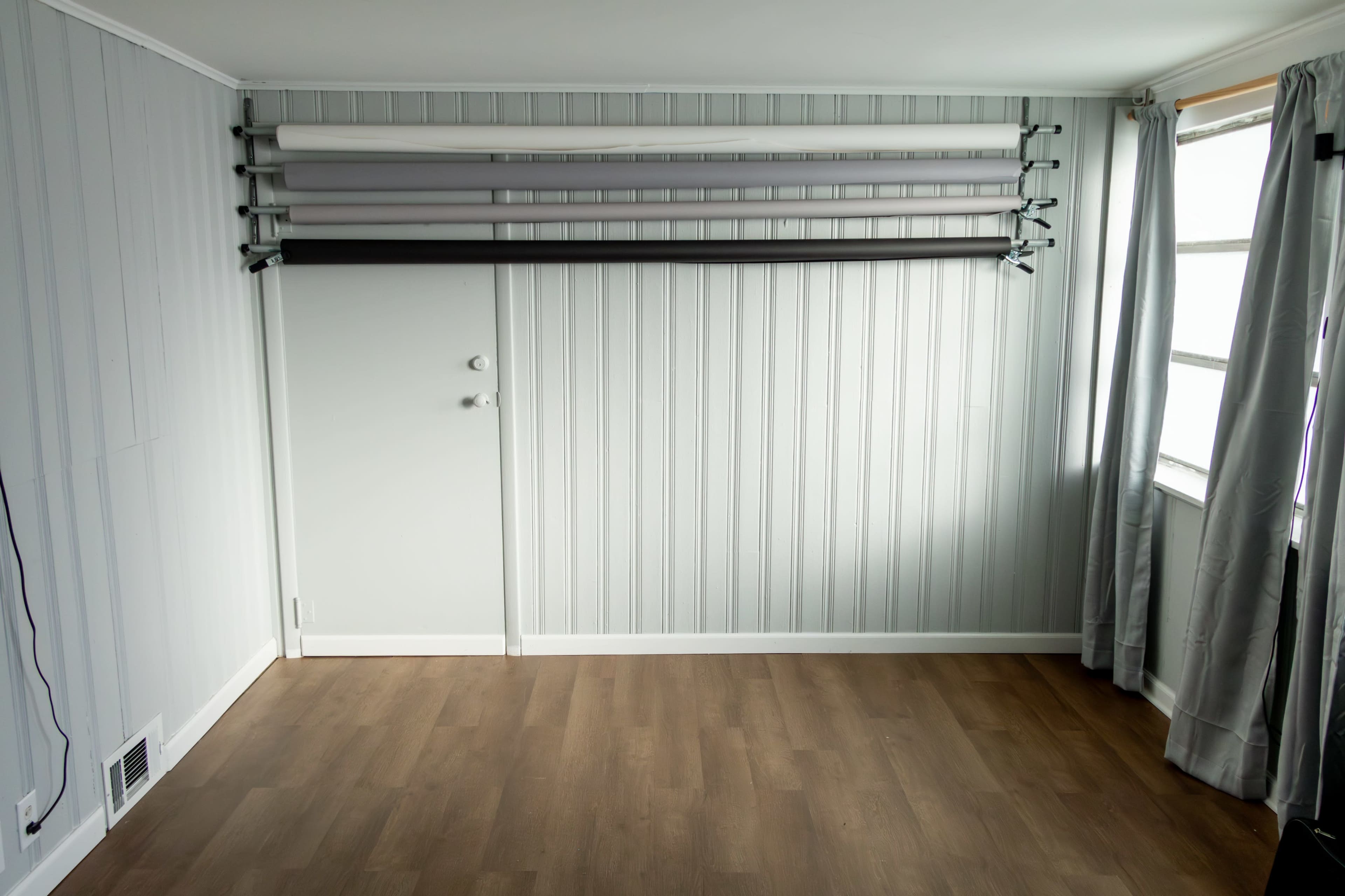 The image shows a minimalist room with wooden flooring, grey paneling on the walls, and several rolled-up fabric backdrops mounted on a rack above a closed door.