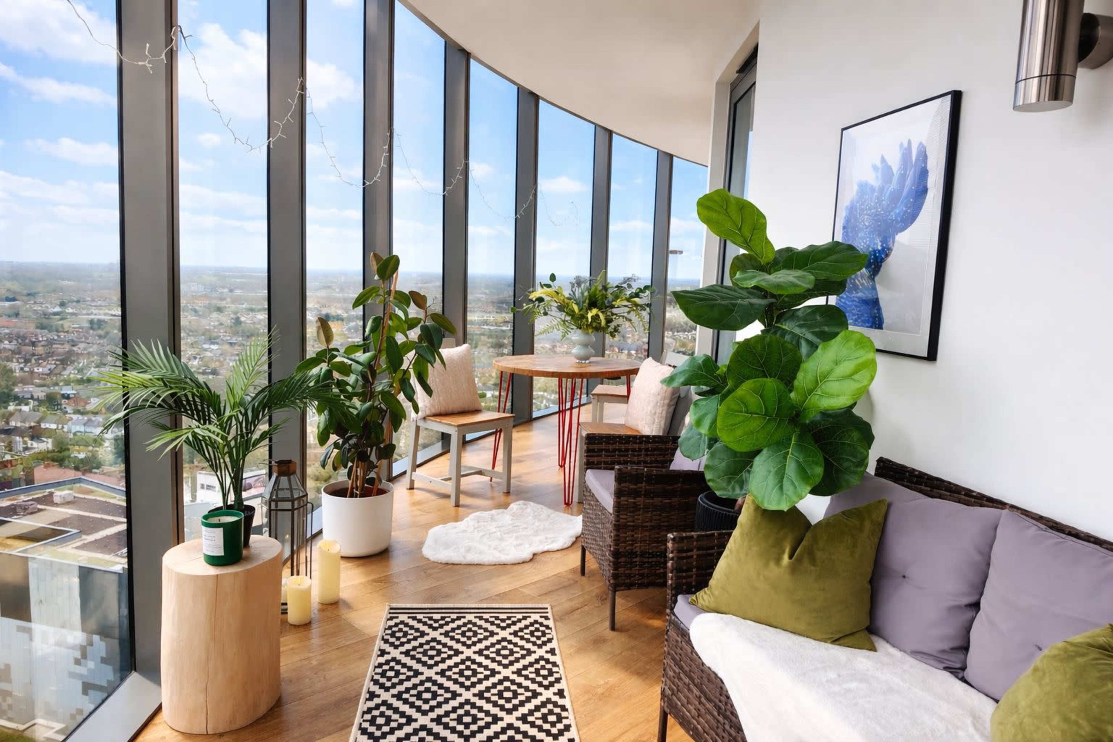 The image shows a sunlit balcony with large glass windows, featuring potted plants, modern furniture, and a dining table with floral decor.