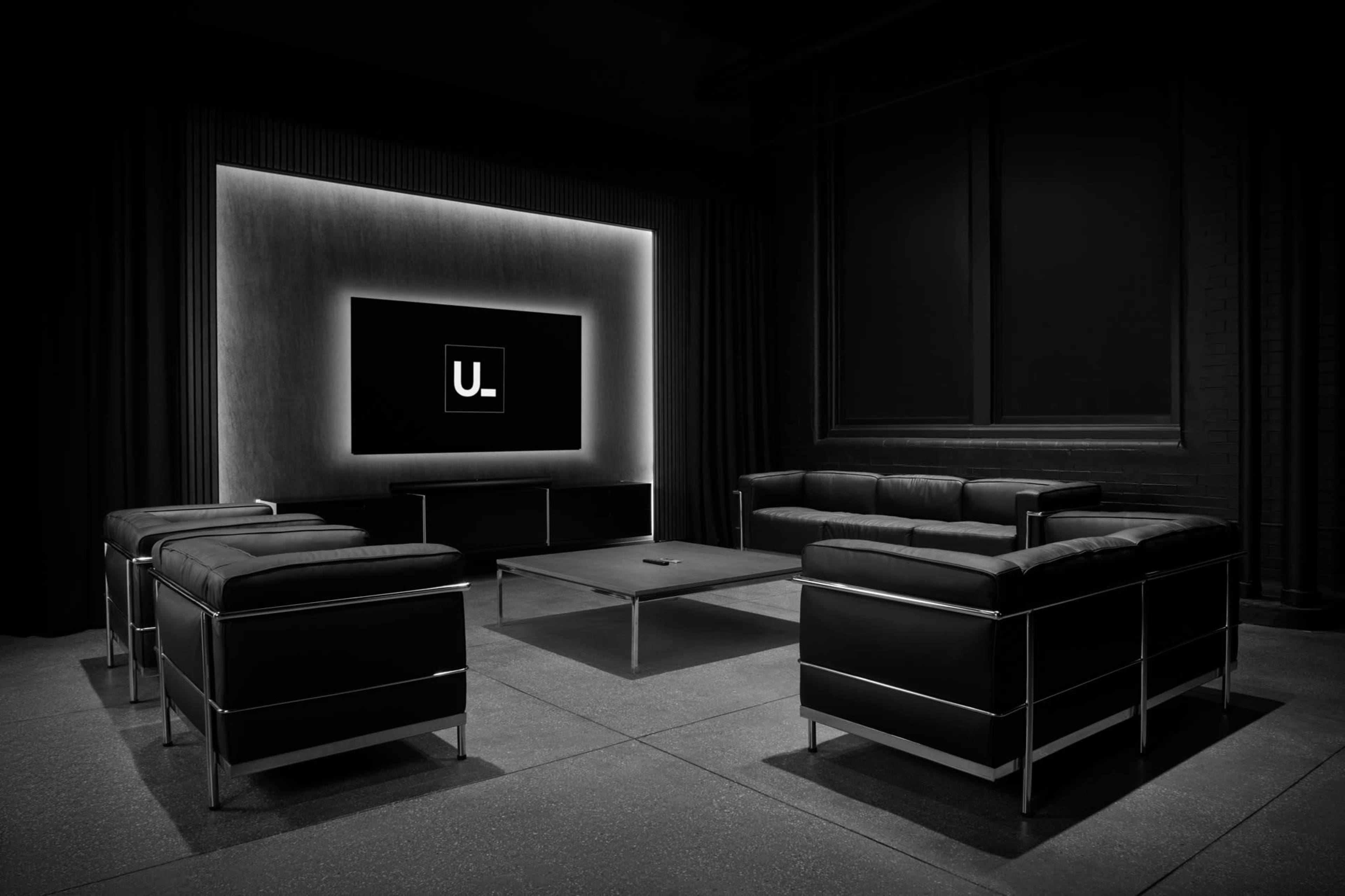 The image depicts a modern, minimalistic lounge area featuring black leather furniture arranged around a glass coffee table, with a lit screen displaying the letter "U." on the wall in a dimly lit room.