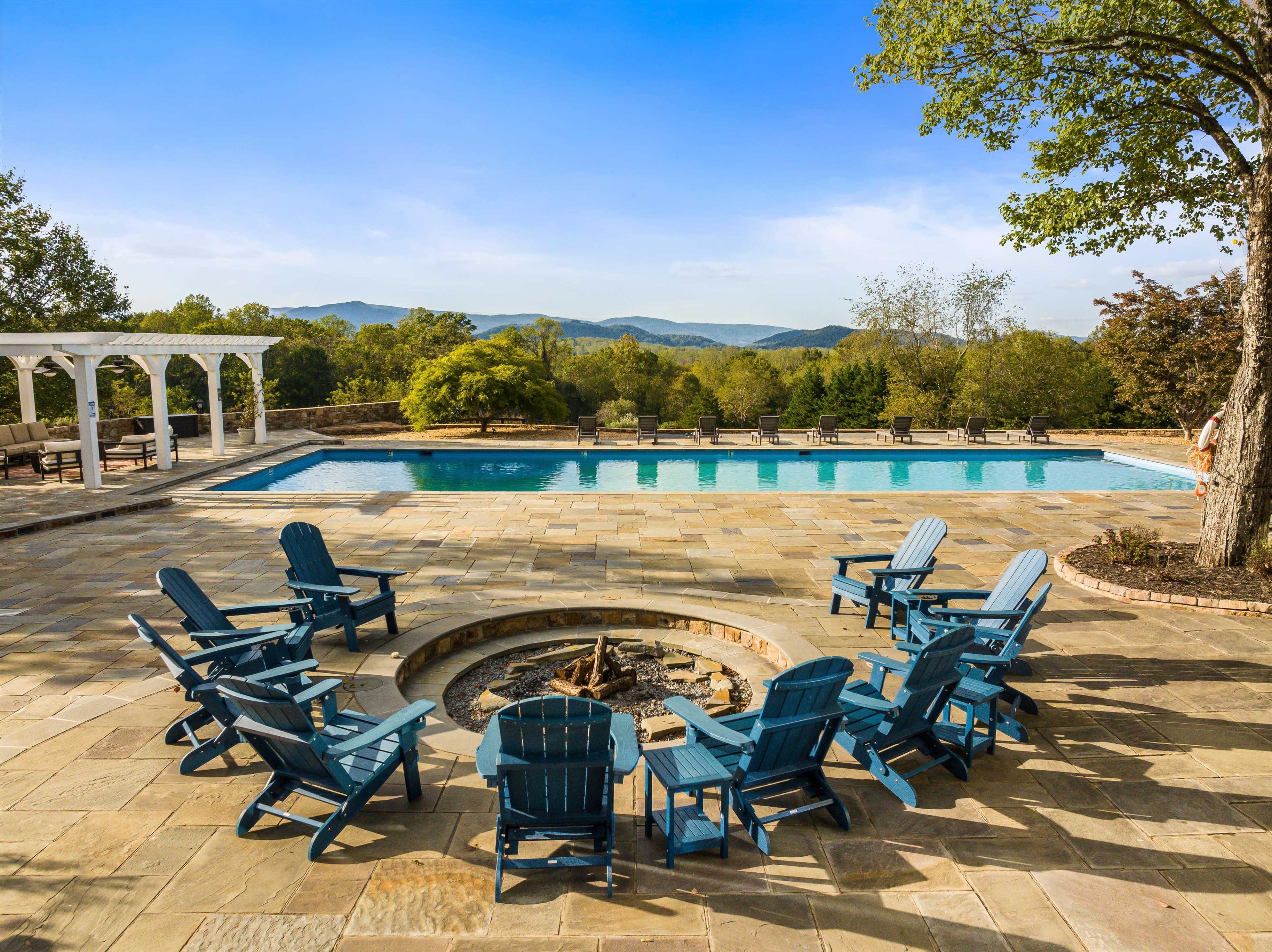 A circular arrangement of blue adirondack chairs surrounds a stone fire pit near a swimming pool with mountains and trees in the background.