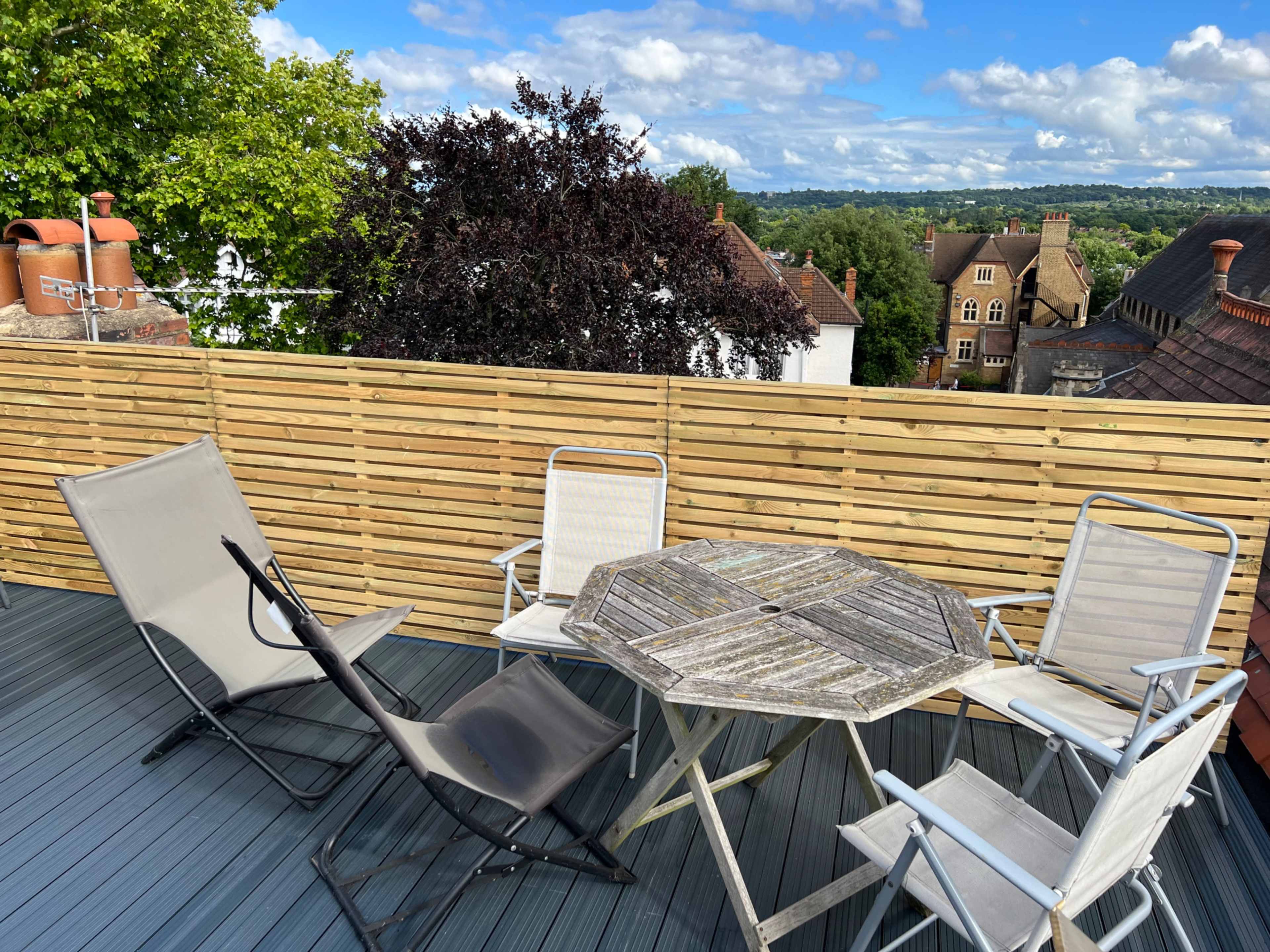 A wooden patio table surrounded by four chairs is set on a deck, overlooking rooftops and trees under a partly cloudy sky.