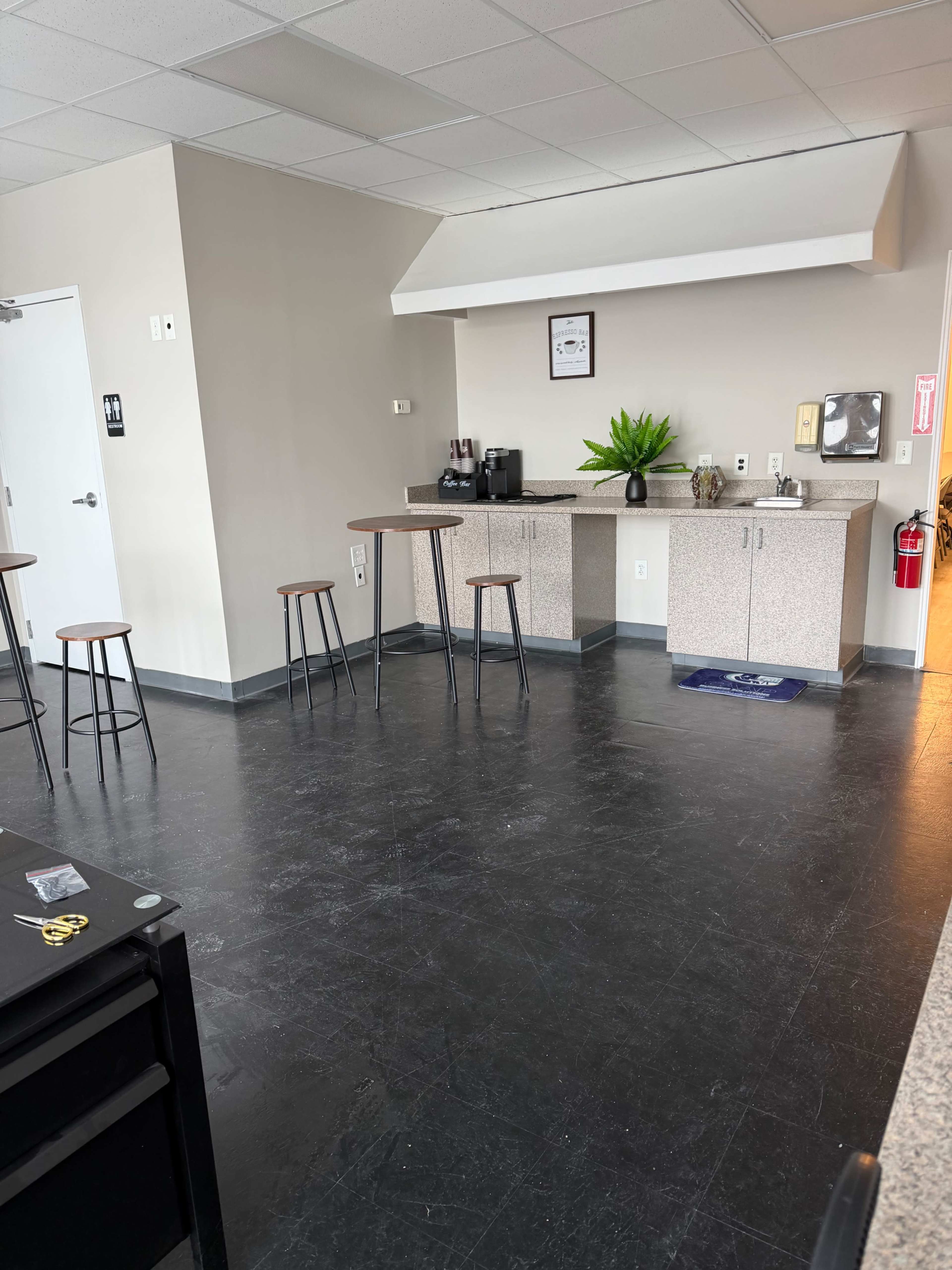 The image shows a break room with a small kitchenette area featuring a countertop, coffee machine, and plants, along with several tall stools around a dark floor.