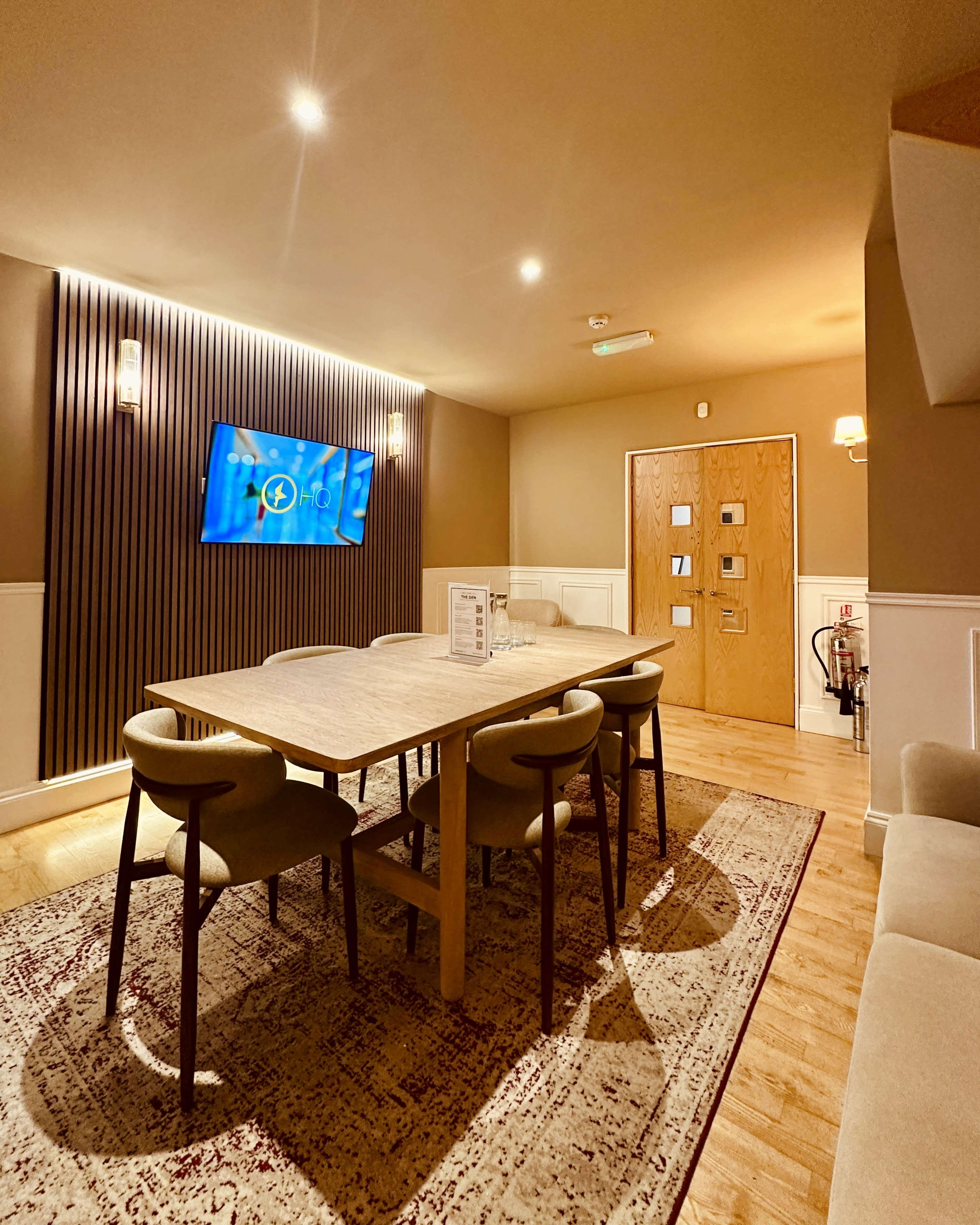The image shows a modern dining area with a wooden table and chairs, a television mounted on the wall, and a decorative rug on the floor.