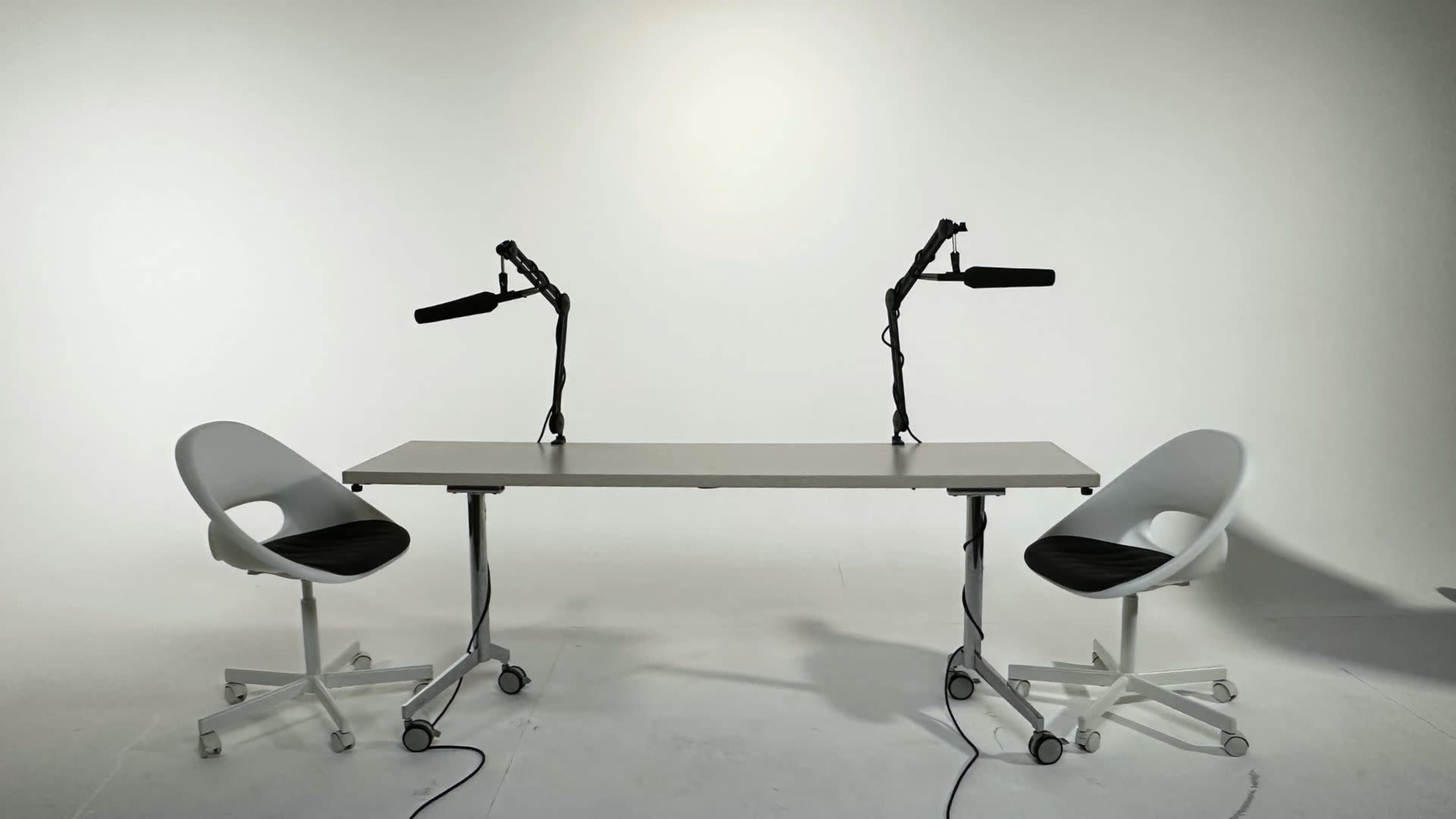 A minimalist studio setup features a central table with two microphones attached, flanked by two wheeled chairs.