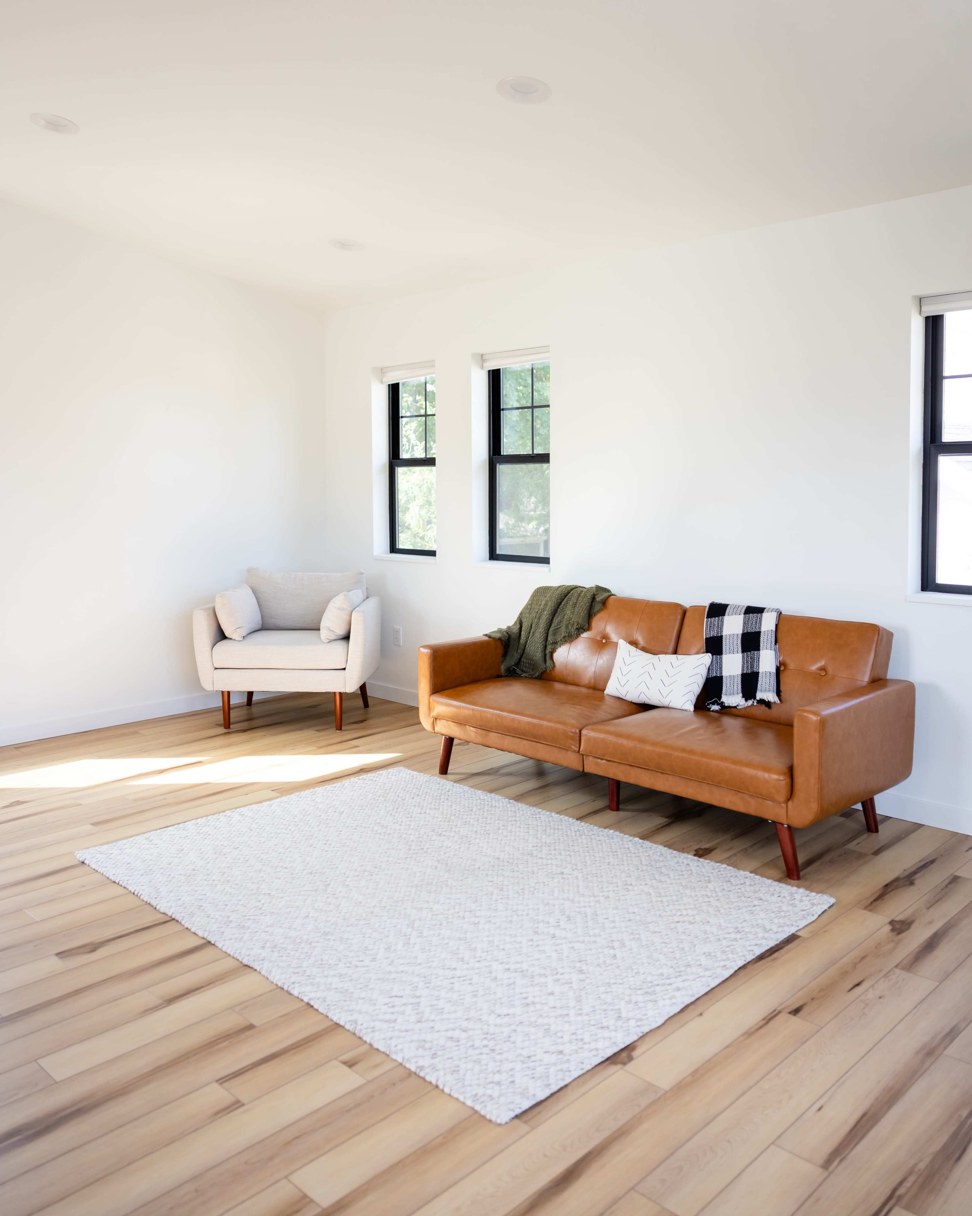 A living room features a brown leather sofa, a light-colored armchair, and a textured area rug on wooden flooring, with large windows allowing natural light.
