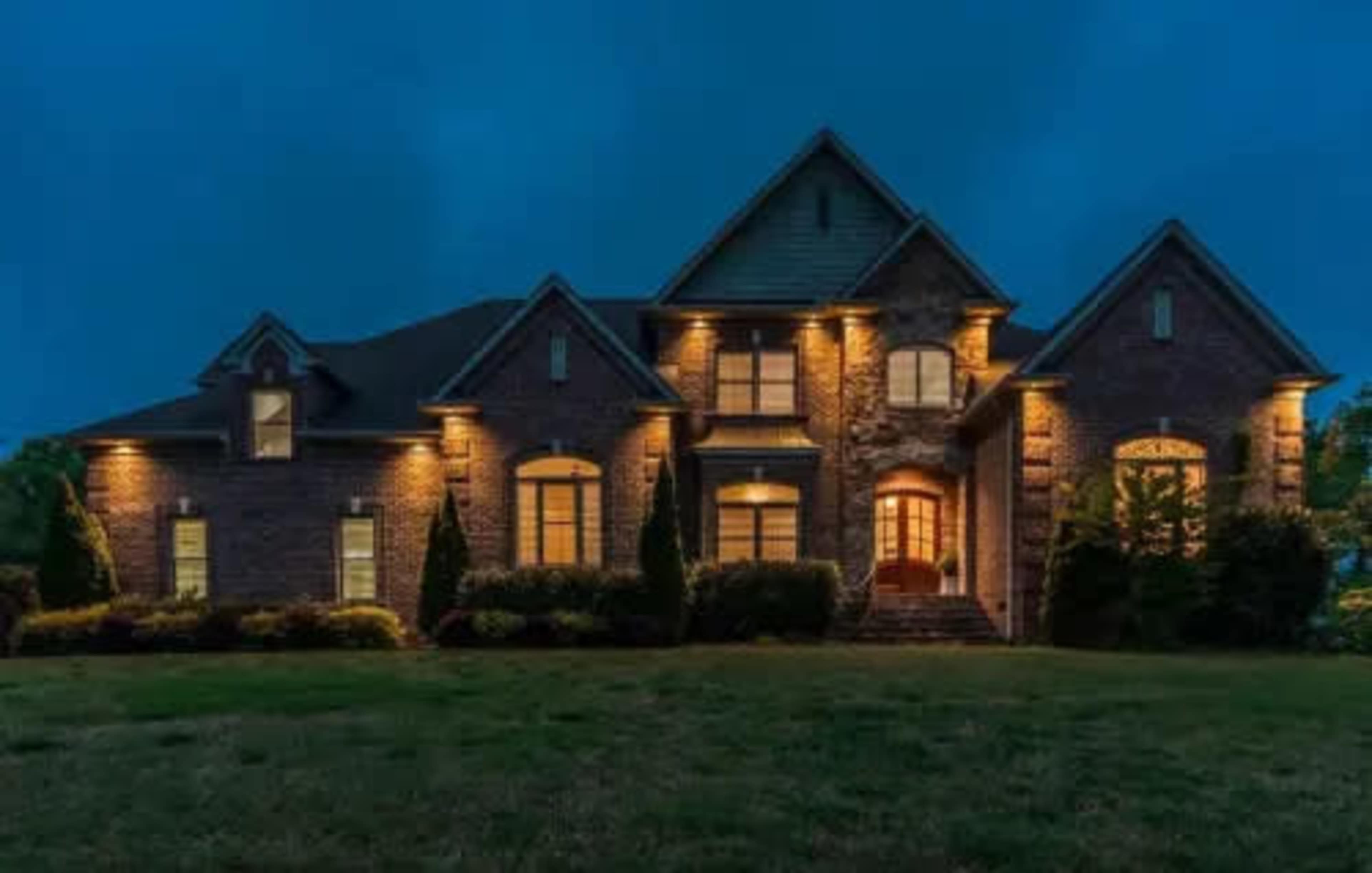 The image shows a large brick house illuminated by exterior lights against a twilight sky.