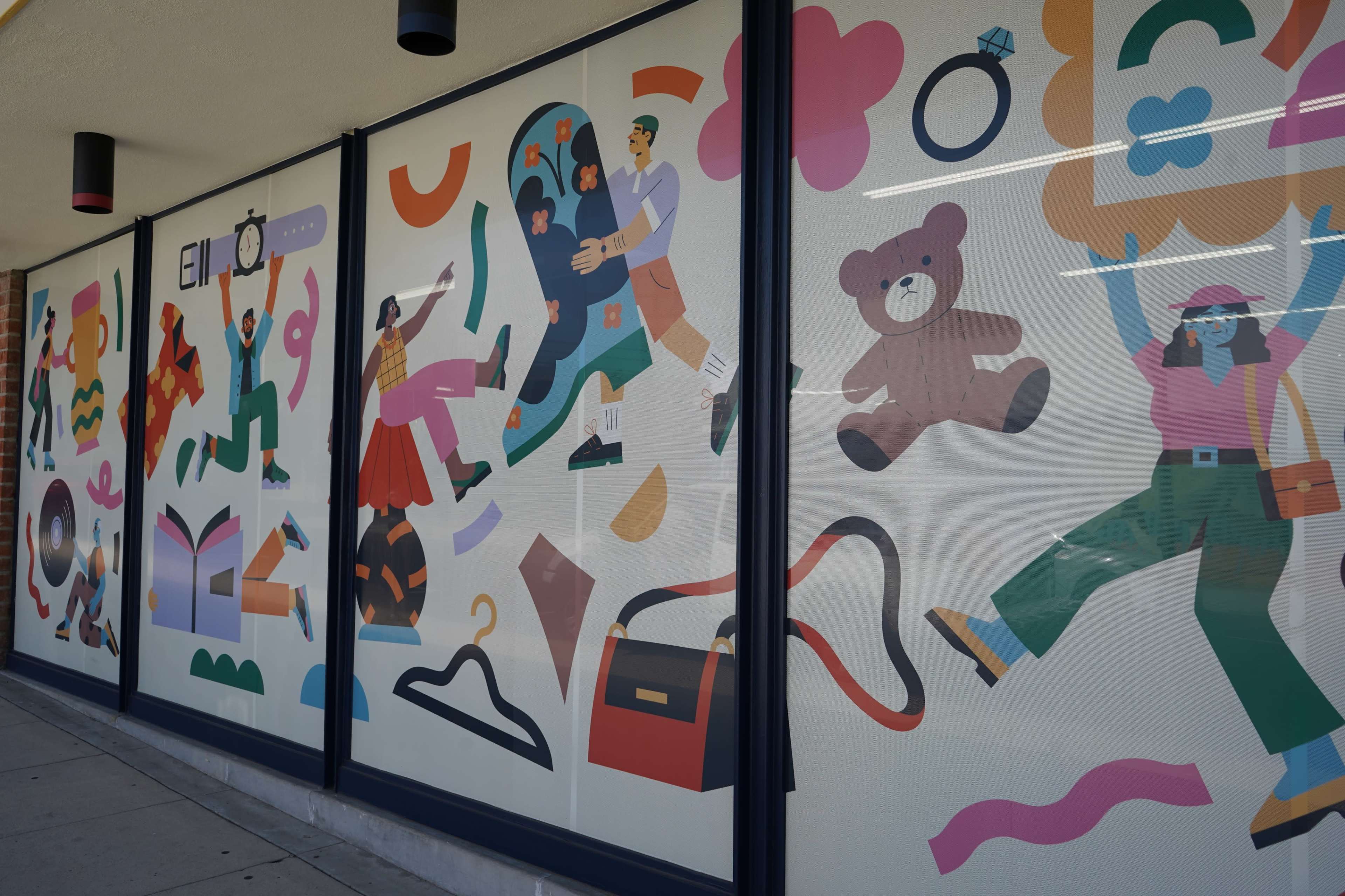 The image shows a colorful mural on a storefront window featuring various illustrations, including people, a bear, a ring, and abstract shapes.