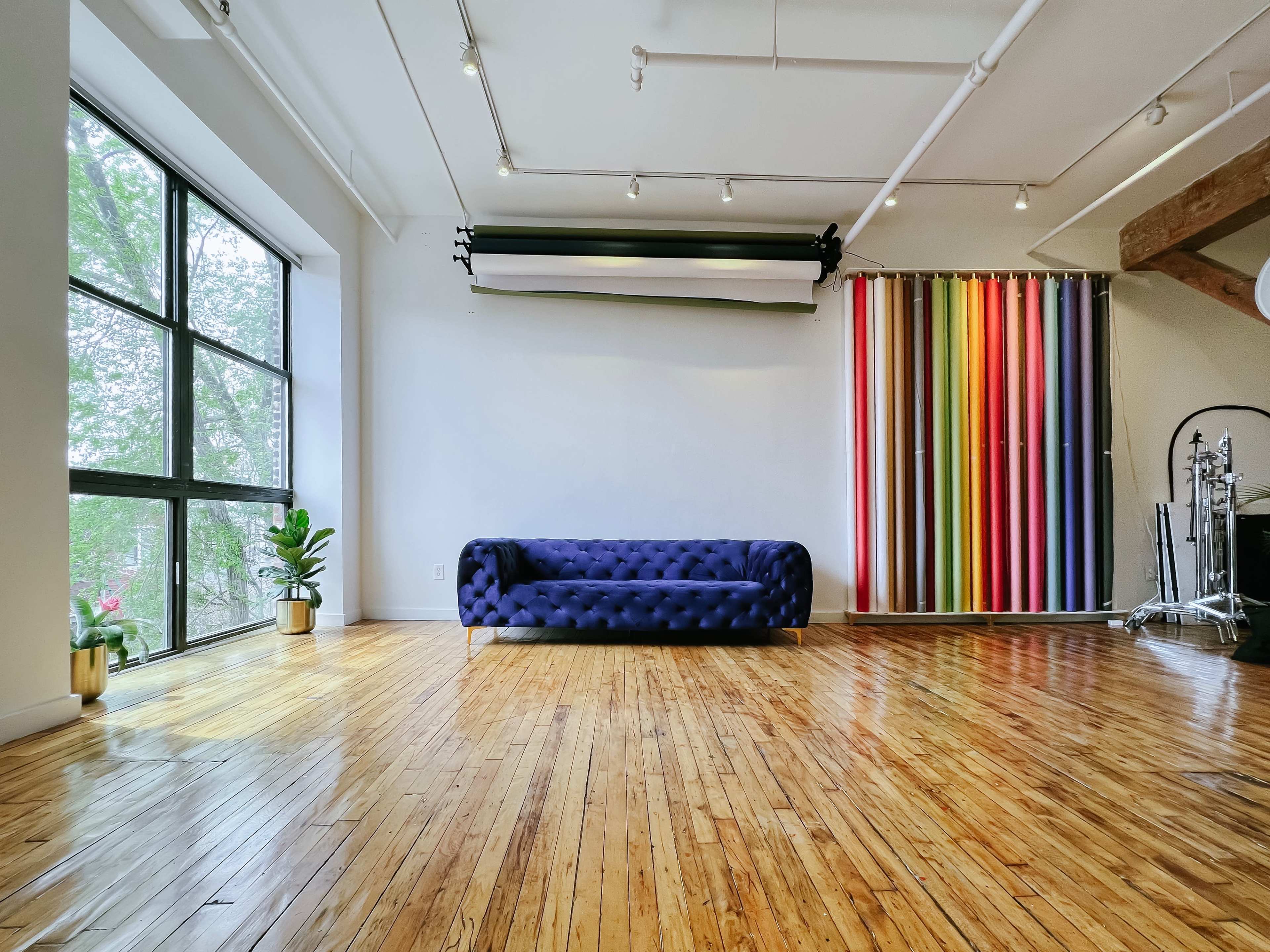 A spacious room with a blue tufted sofa placed against a wall, large windows letting in natural light, and a colorful striped backdrop on one side.
