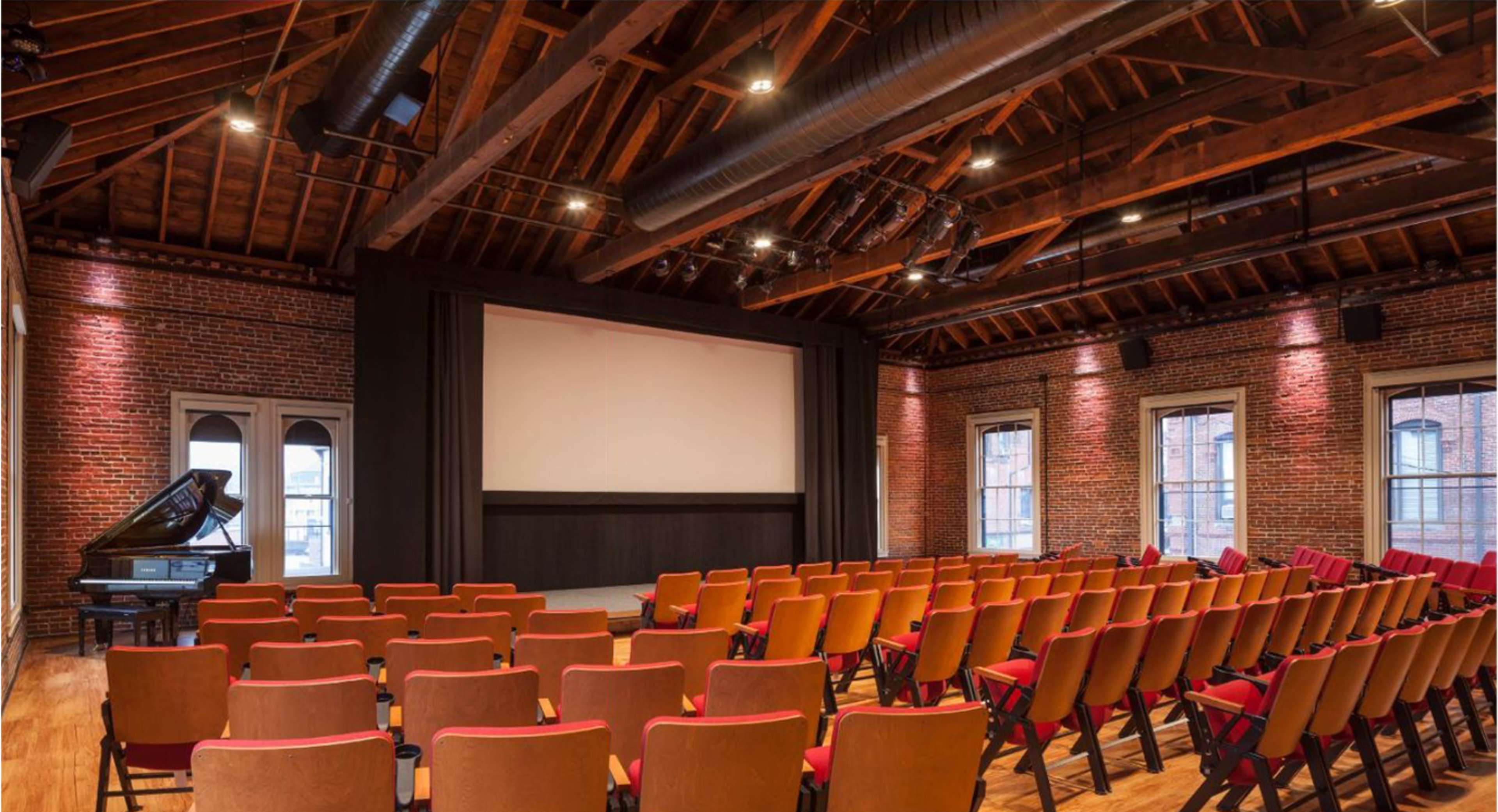 An auditorium with wooden beams, red-brick walls, a large screen at the front, and rows of red-covered chairs facing the stage.