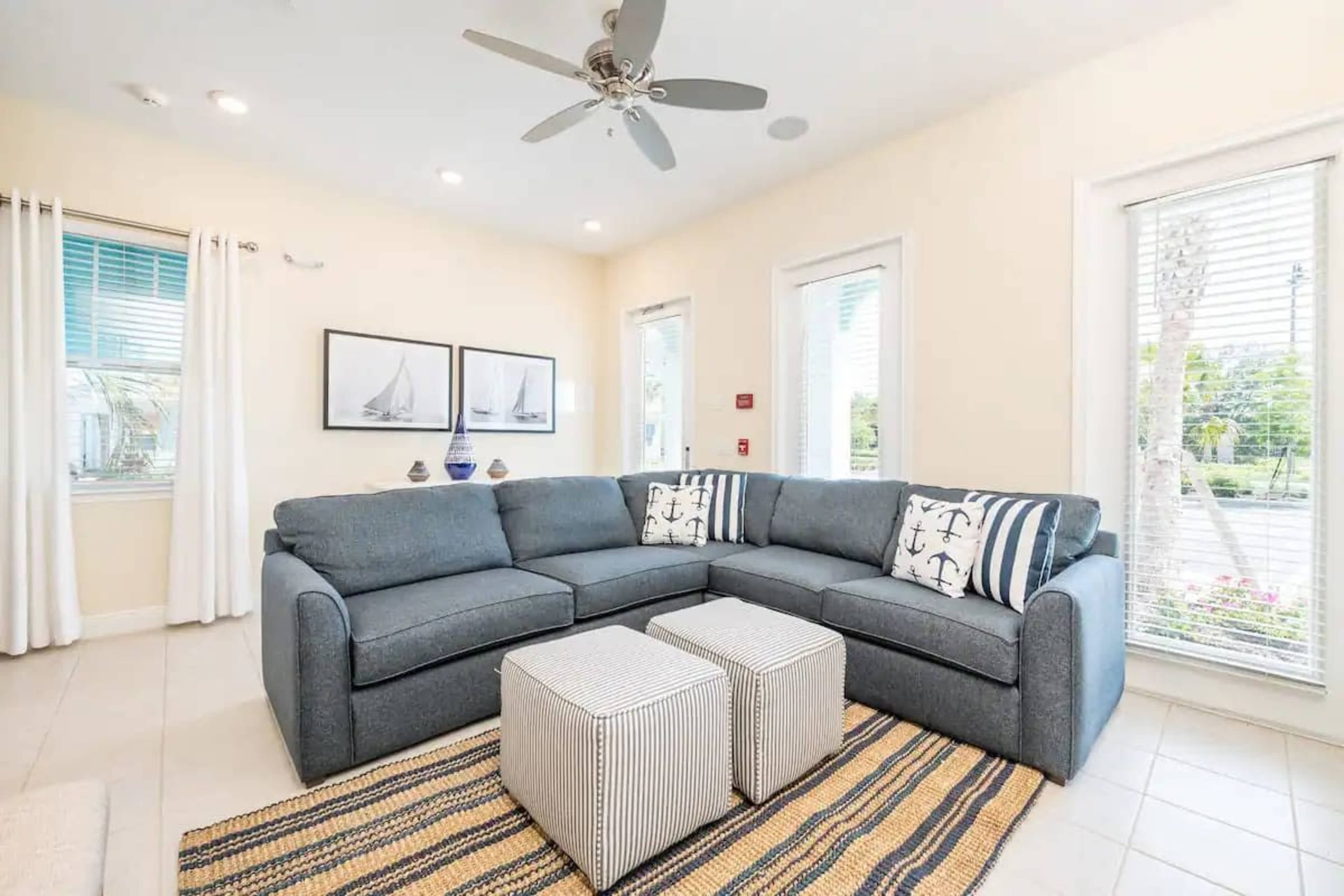 A cozy living room with a gray sectional sofa, two striped ottomans, and windows letting in natural light.