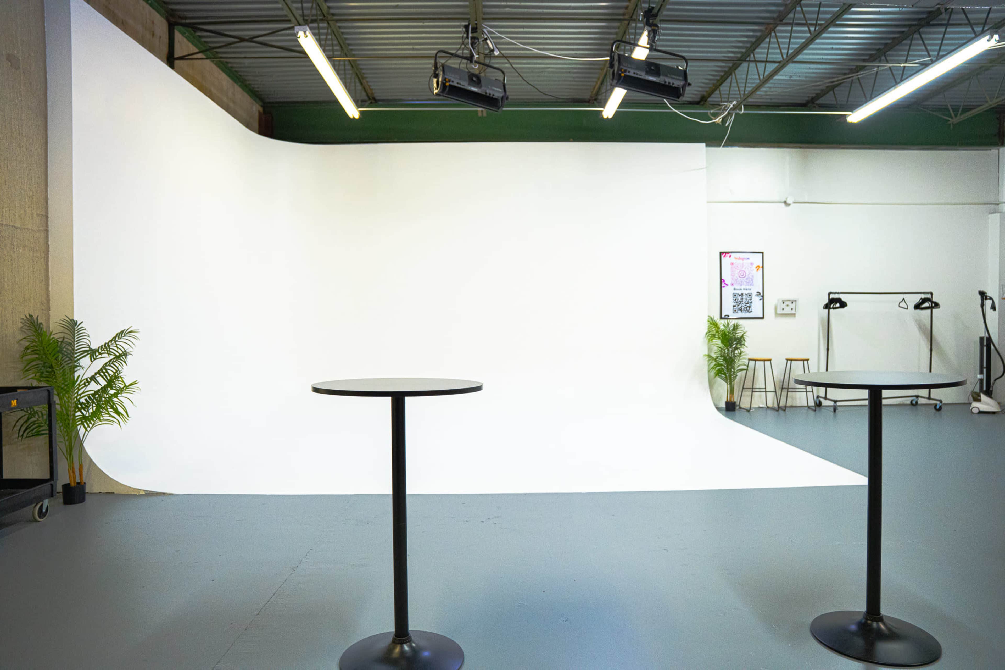 The image shows a bright studio space with two black tables in front of a large white backdrop, illuminated by overhead lights.