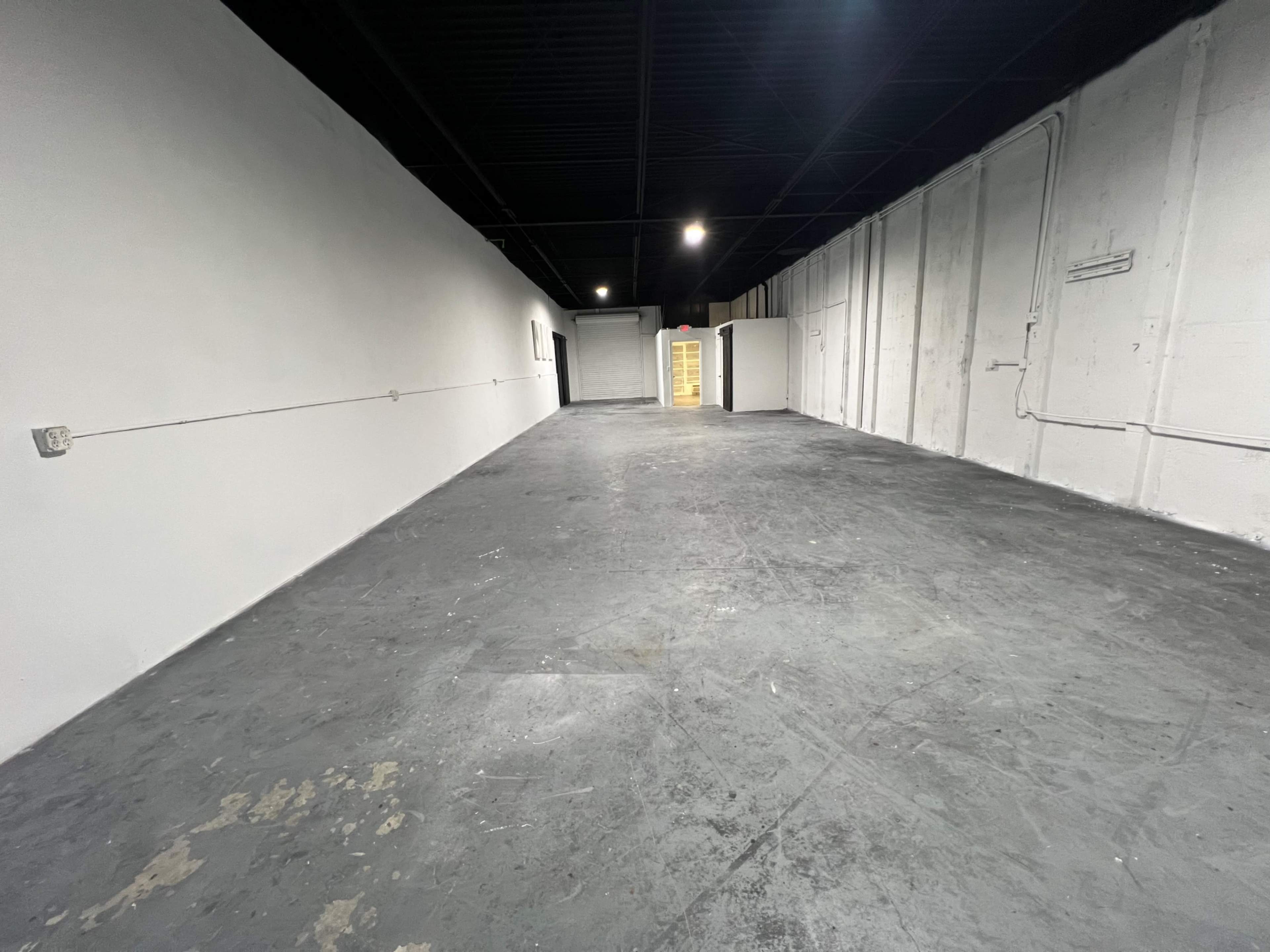 The image shows an empty warehouse space with a gray floor, whitewashed walls, and overhead lighting along the ceiling.