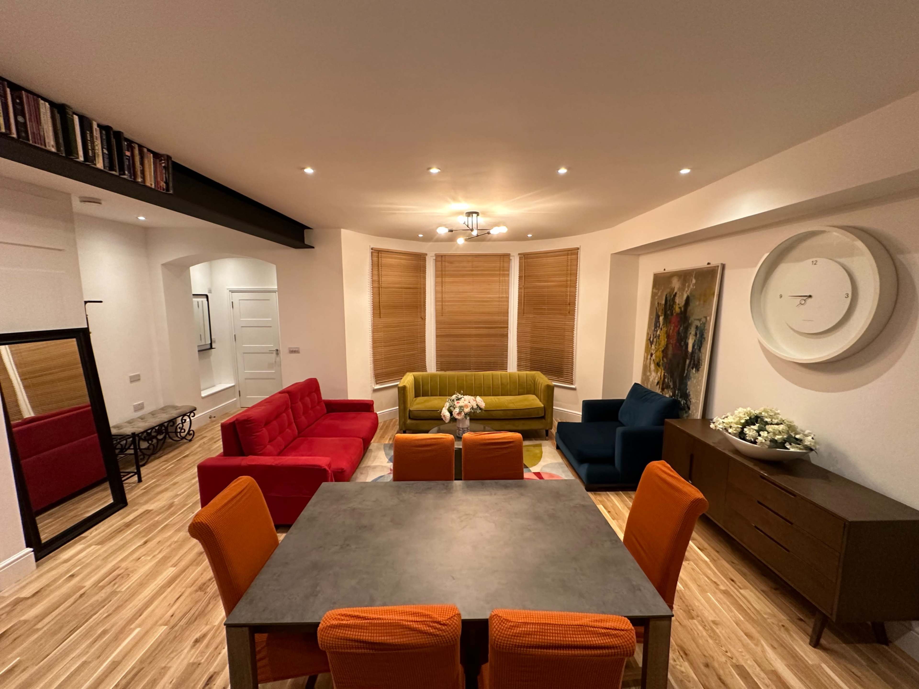 A modern living space features a dining table surrounded by orange chairs, three sofas in red, green, and blue, and large windows with wooden blinds.