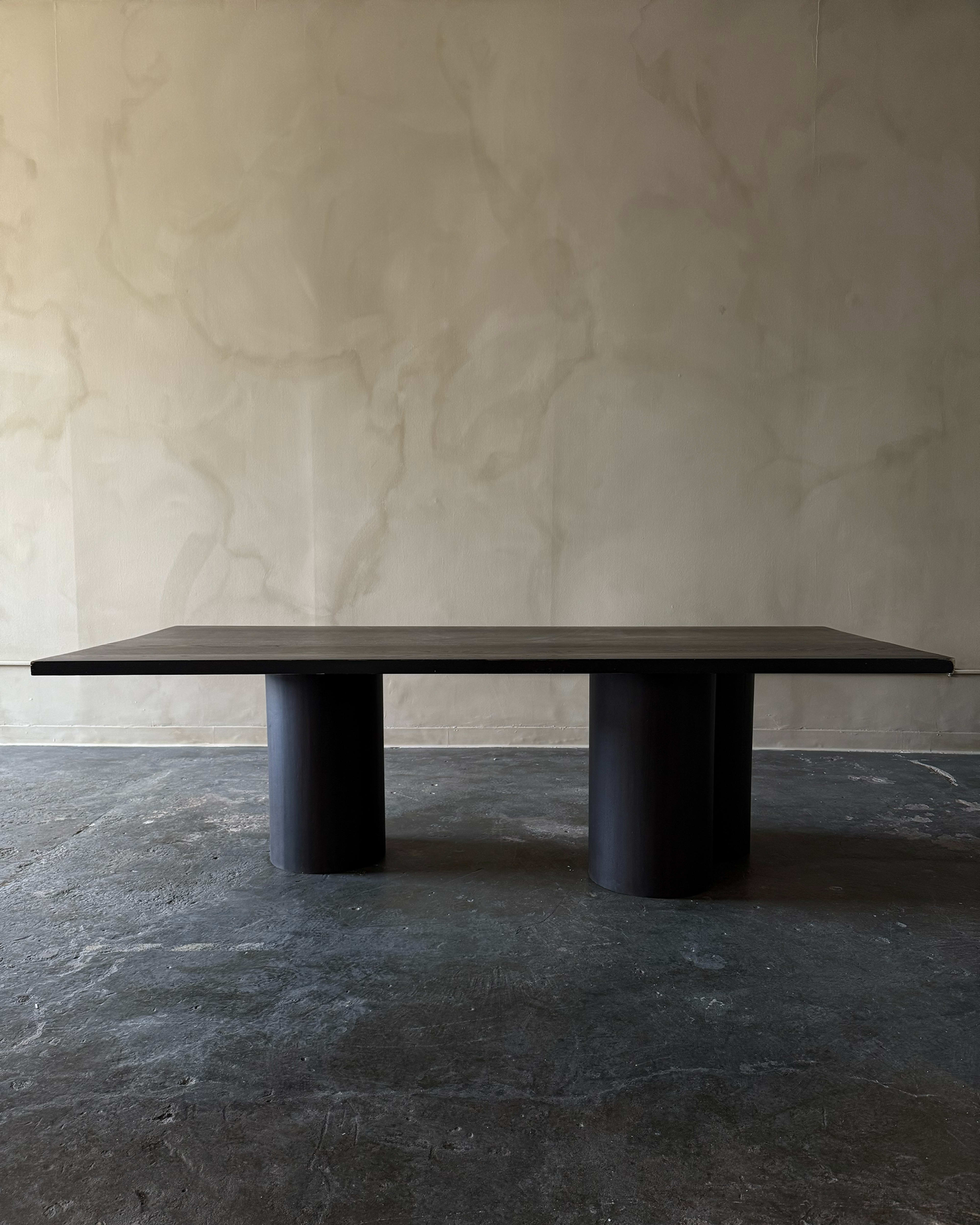 A large, dark wooden table with a flat top is supported by two round columns, set against a textured wall.