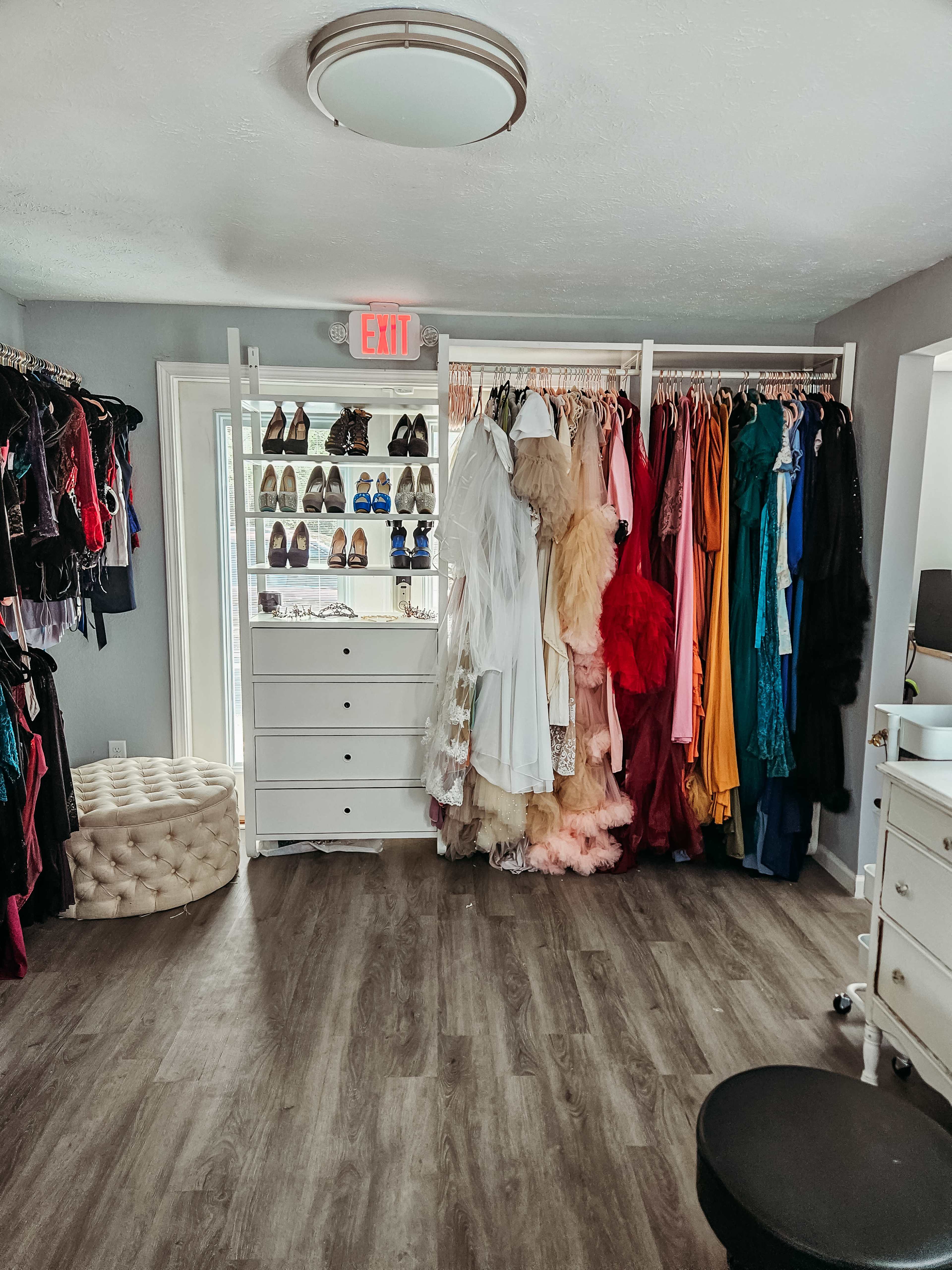 A brightly lit closet with a variety of colorful dresses hanging on racks, shelves of shoes, a tufted ottoman, and a white dresser.