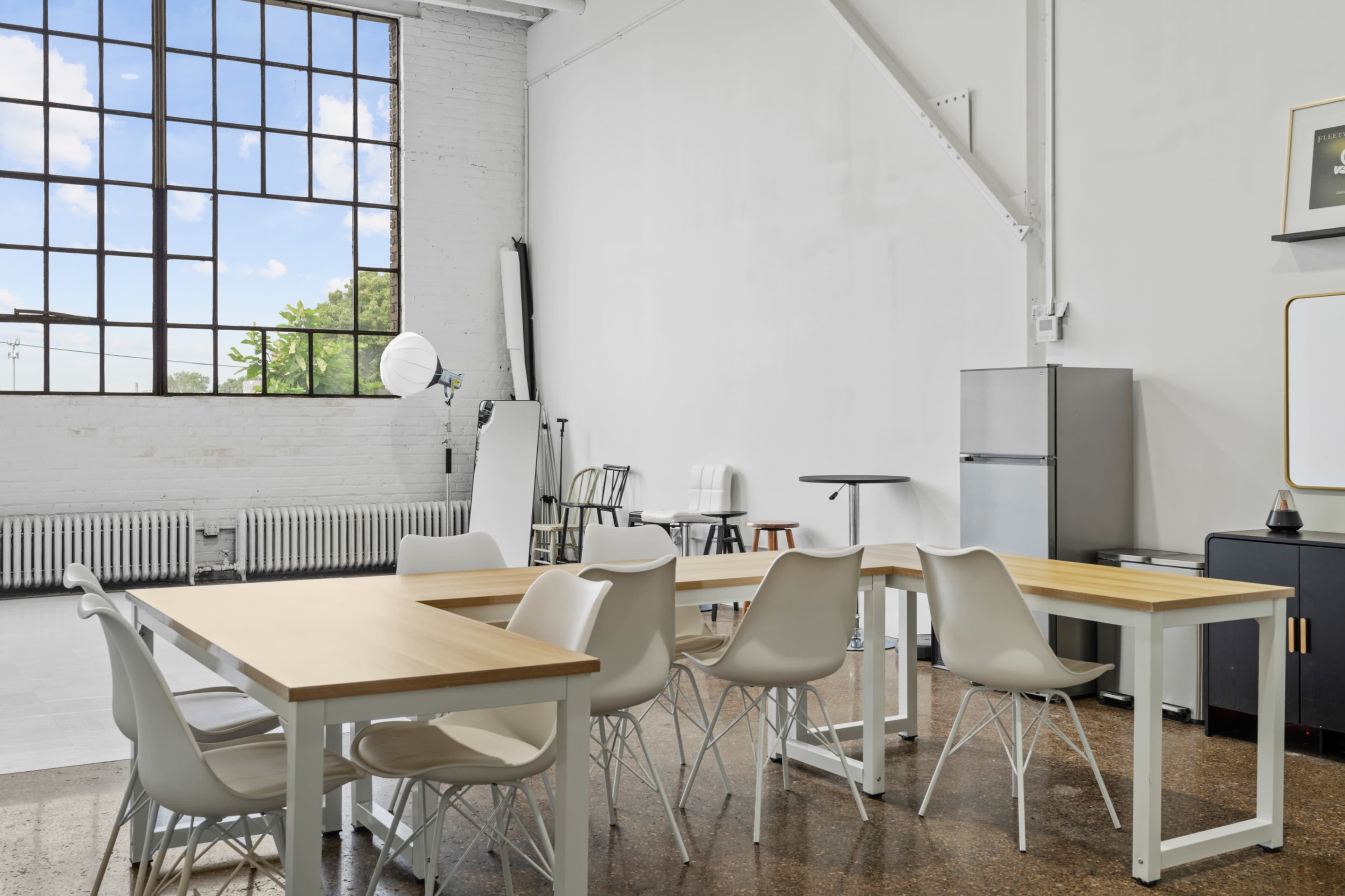 The image shows a brightly lit interior space with several wooden tables and modern white chairs, large windows, and a minimalistic design featuring a refrigerator and a black cabinet.
