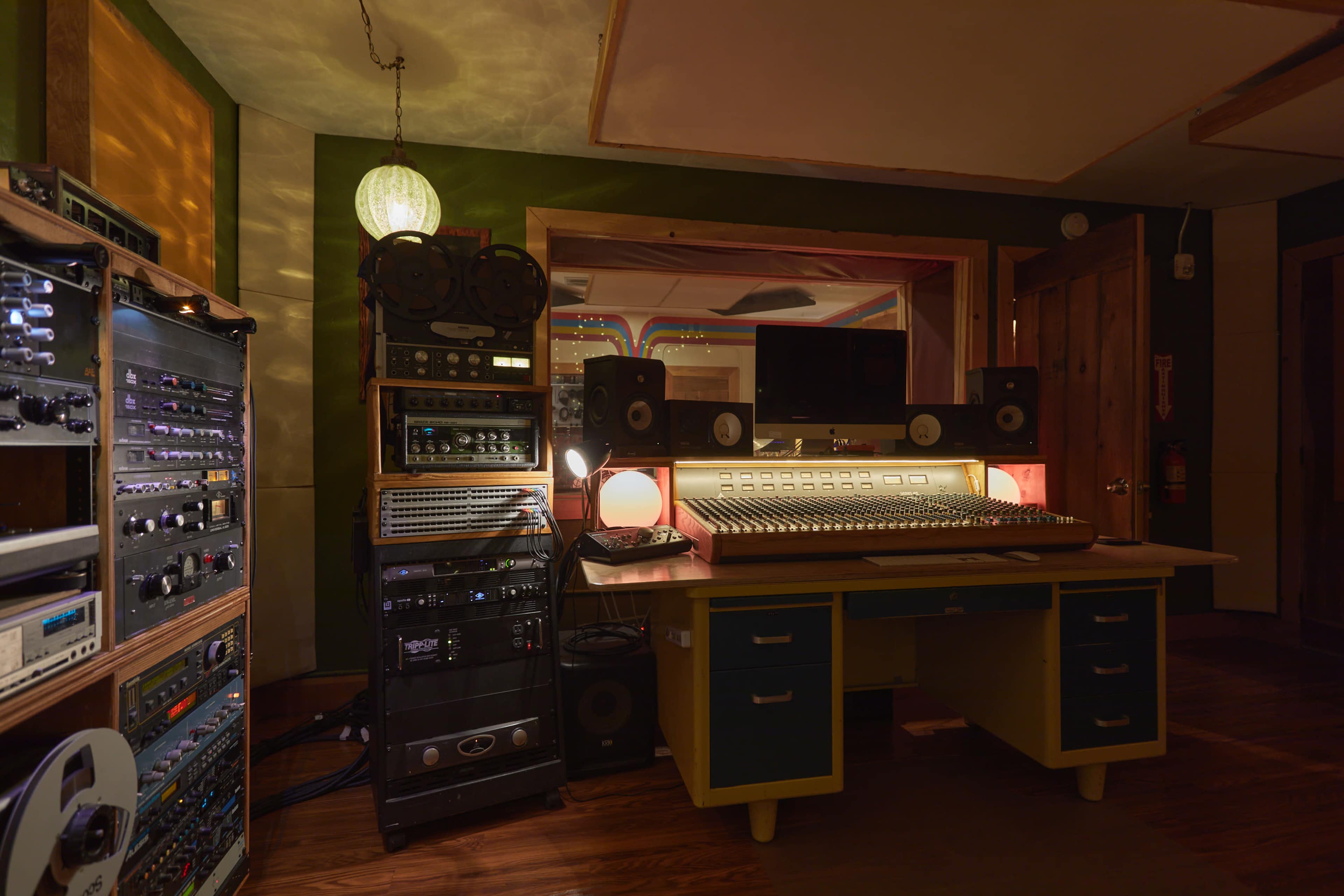 The image shows a dimly lit music recording studio with audio equipment, a mixing console, and speakers arranged around a central desk.