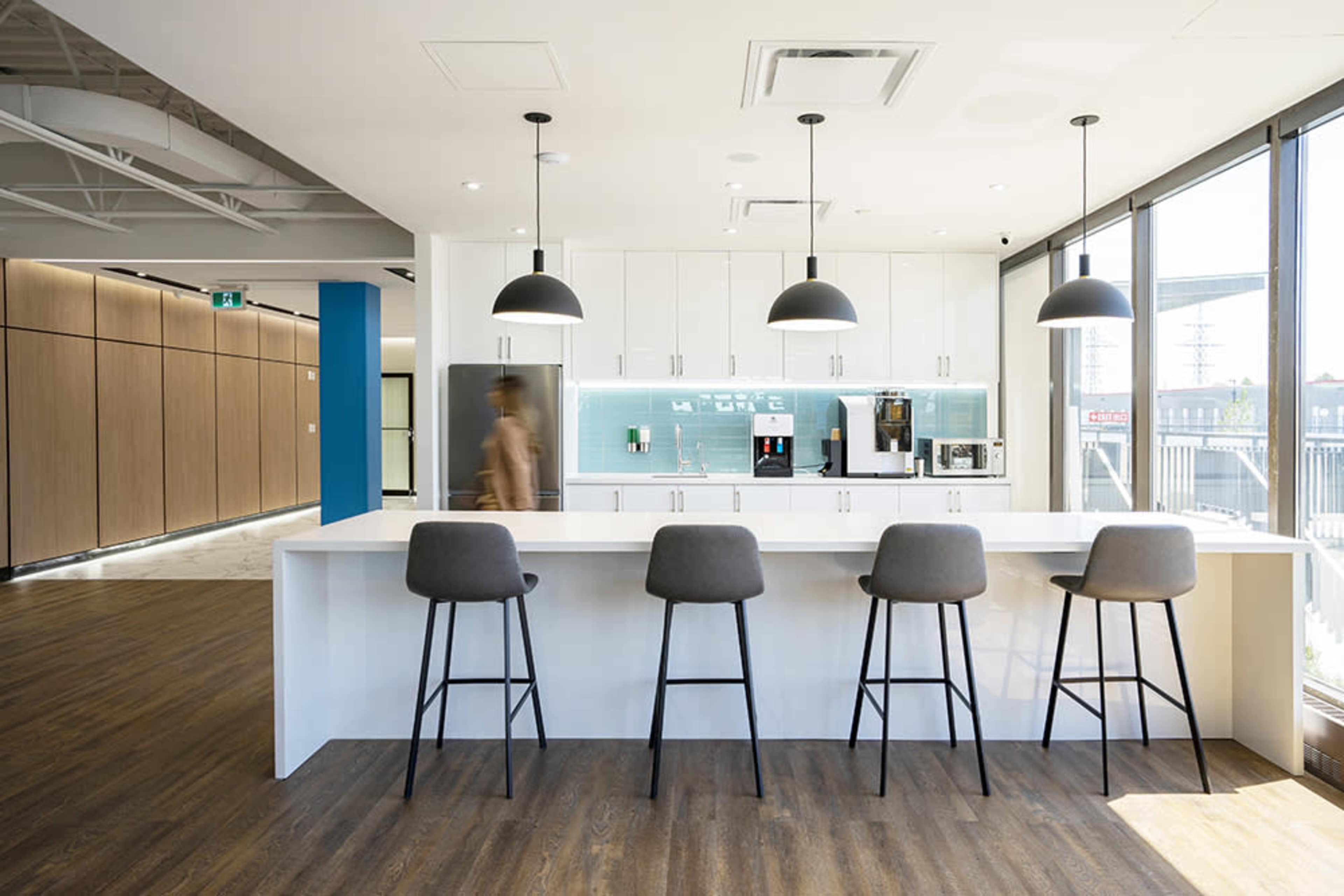 A modern kitchen with a white countertop and four bar stools, featuring a coffee machine and other appliances against a light blue backsplash.
