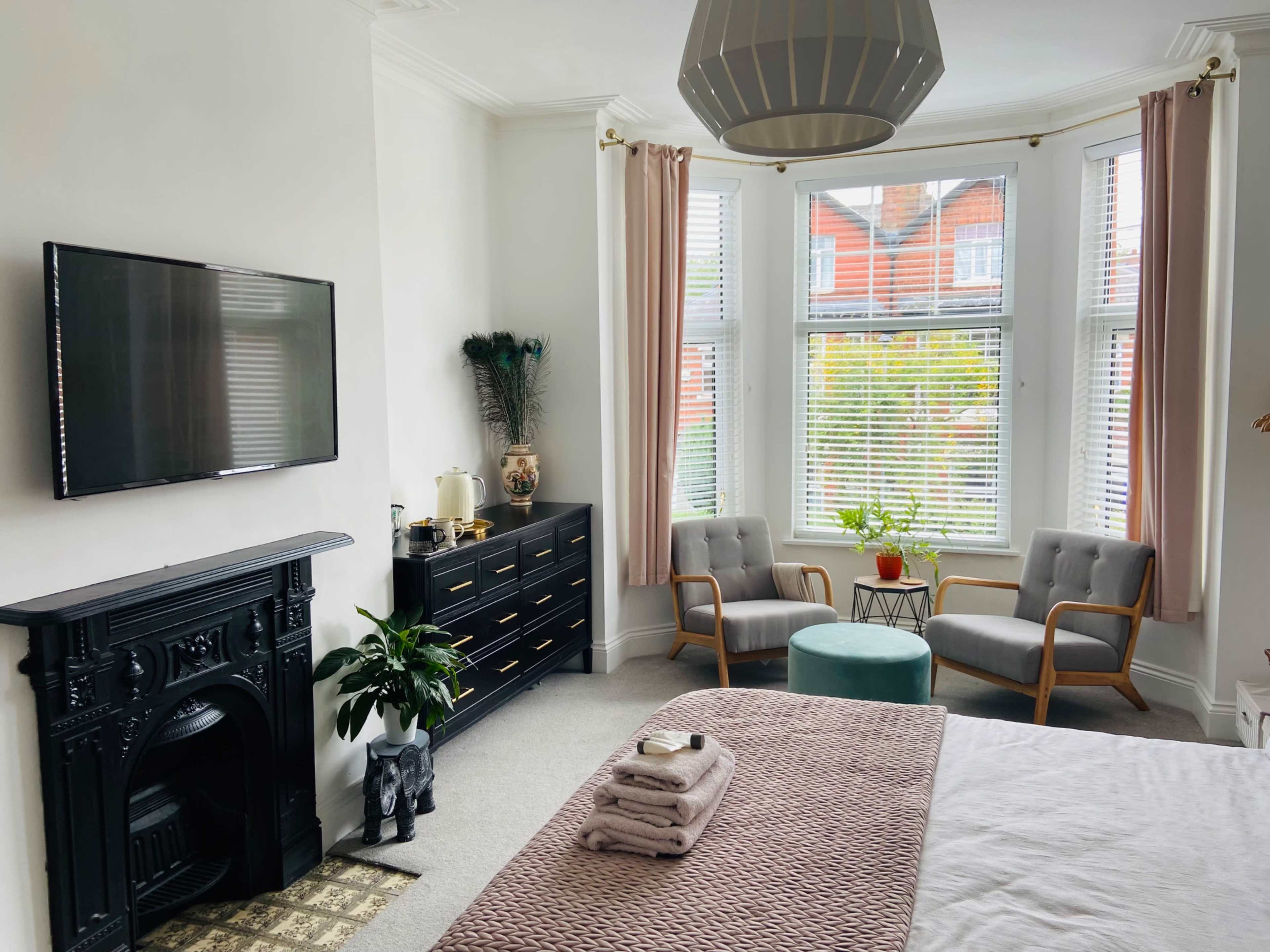 A furnished bedroom features a bed with neatly stacked towels, a television mounted on the wall, and two armchairs near a window, accompanied by decorative plants.