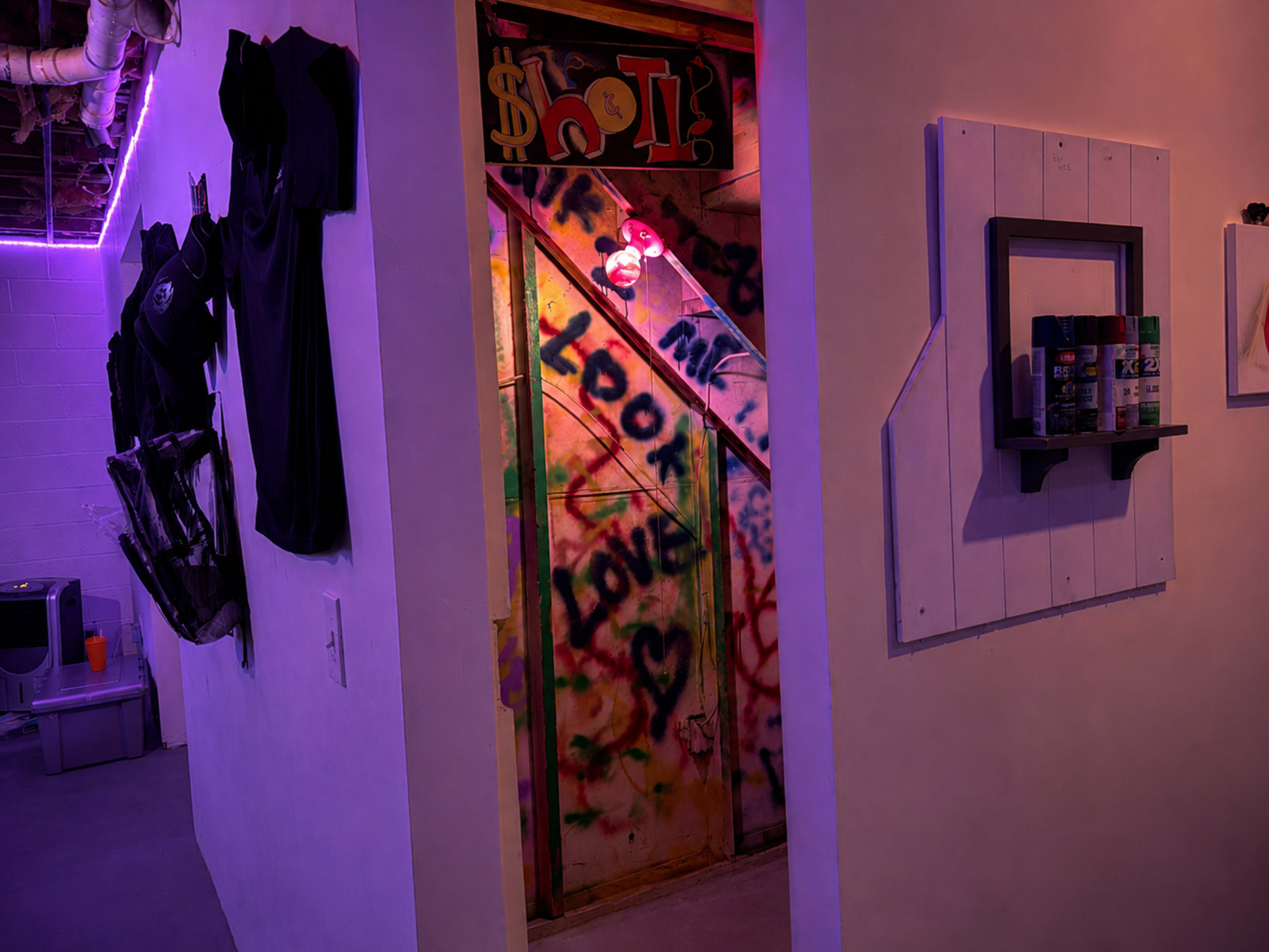 A dimly lit hallway features a colorful graffiti-covered door leading to a staircase, with black clothing hanging on the walls.
