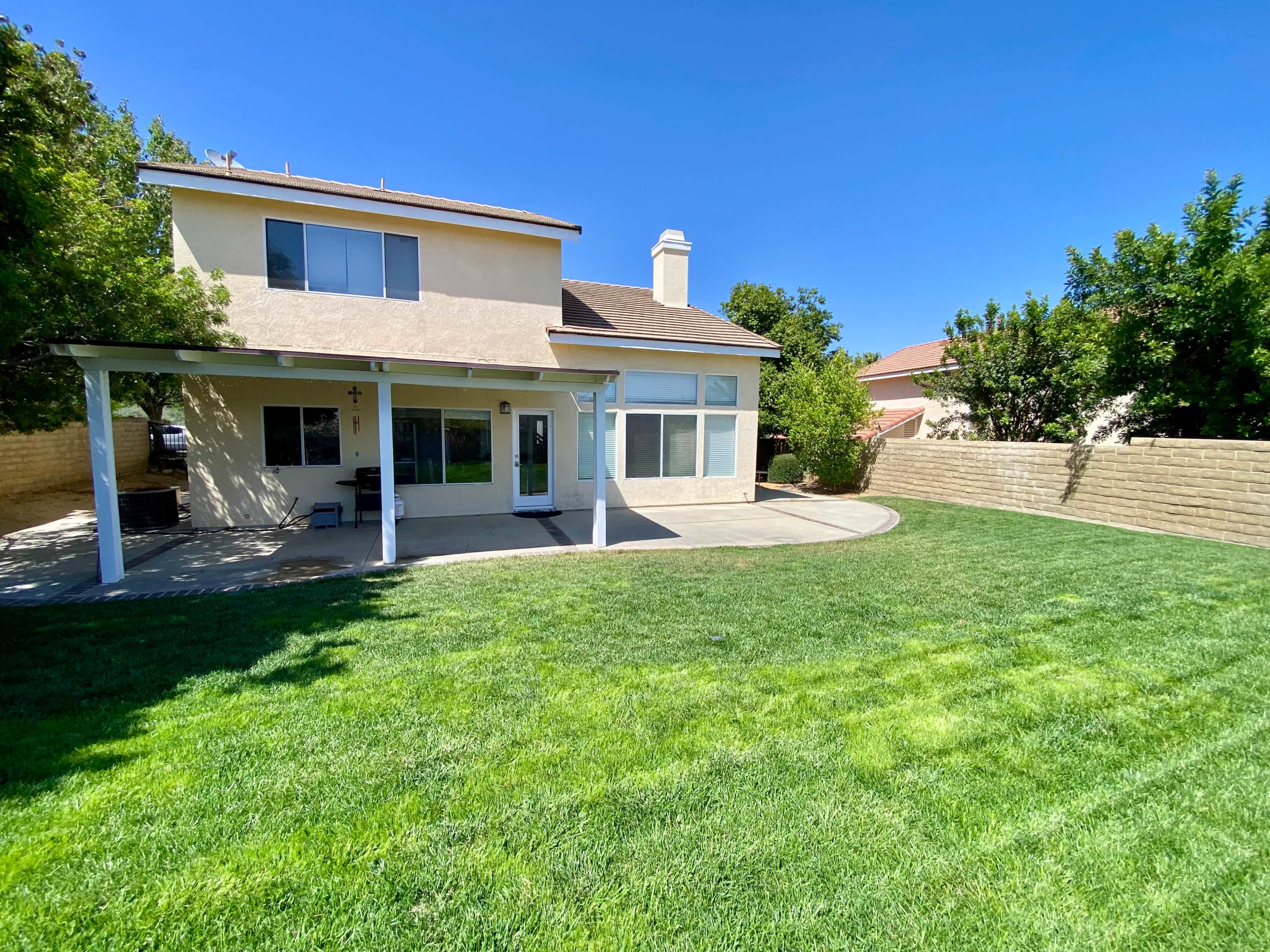The image shows a two-story house with a patio in the backyard, surrounded by green grass and tall shrubs.