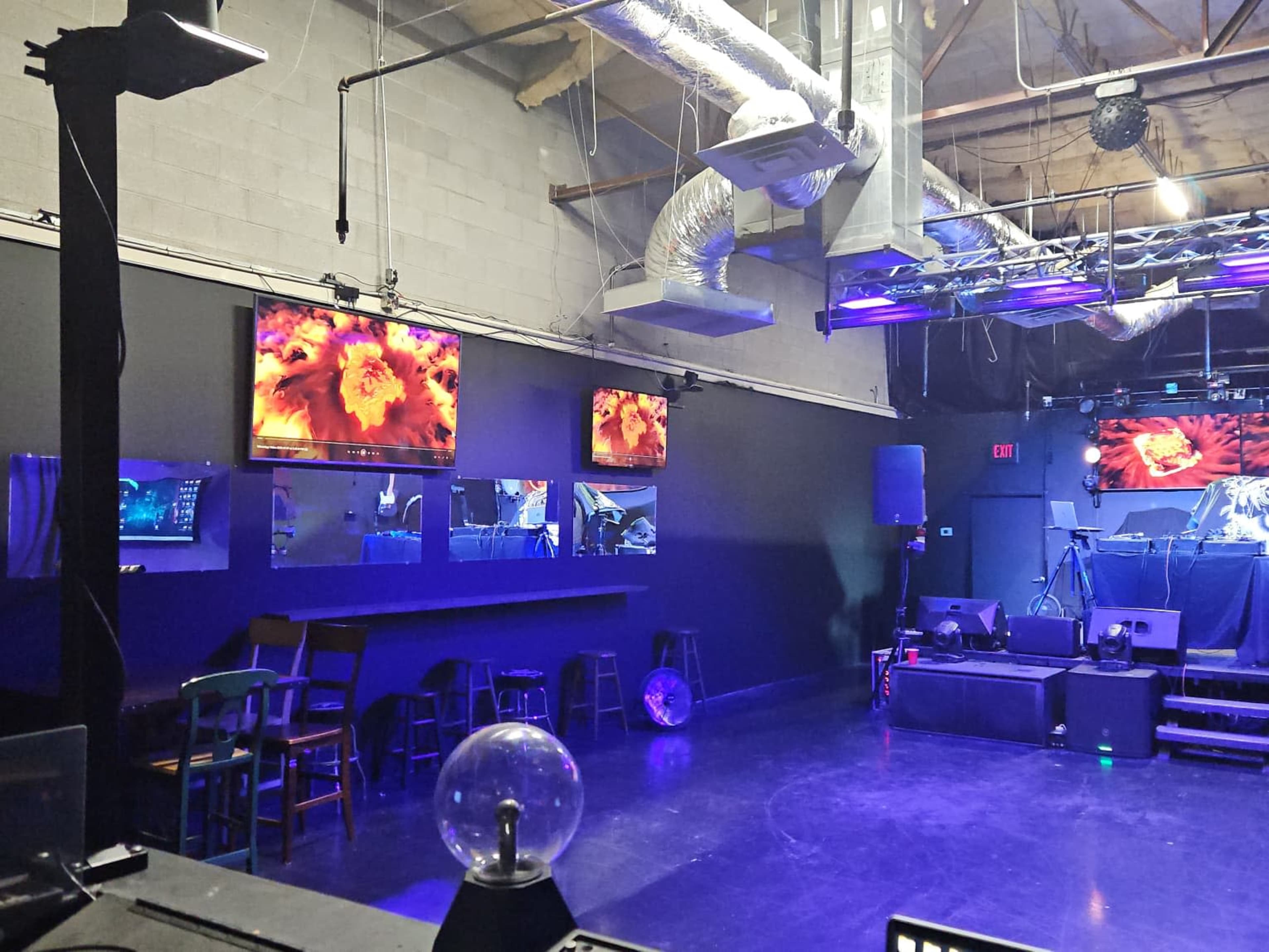 The image shows a dimly lit music venue with multiple screens displaying visuals, a performance stage, and seating arrangements against a black wall.