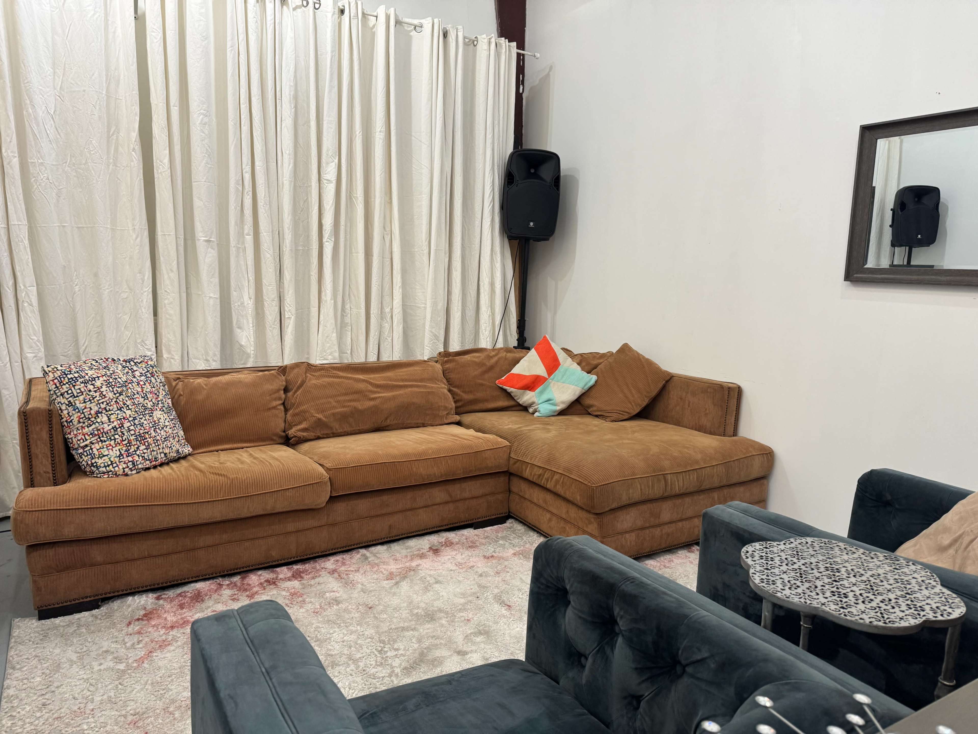 The image depicts a modern living room setup with a brown sectional sofa, two teal chairs, a small decorative table, and light-colored curtains.