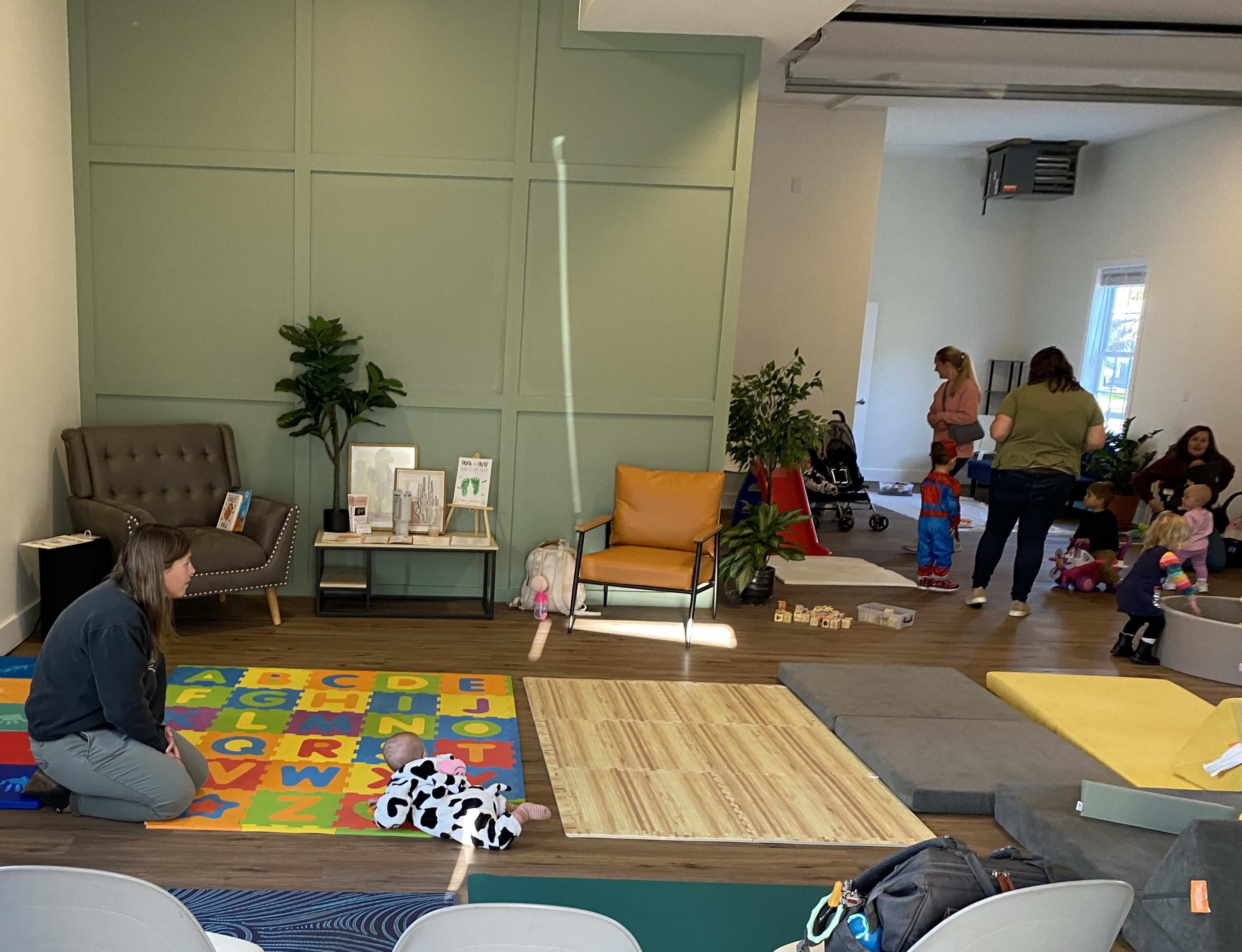 A playroom features colorful alphabet mats, a gray armchair, and several children engaged in activities while adults supervise.
