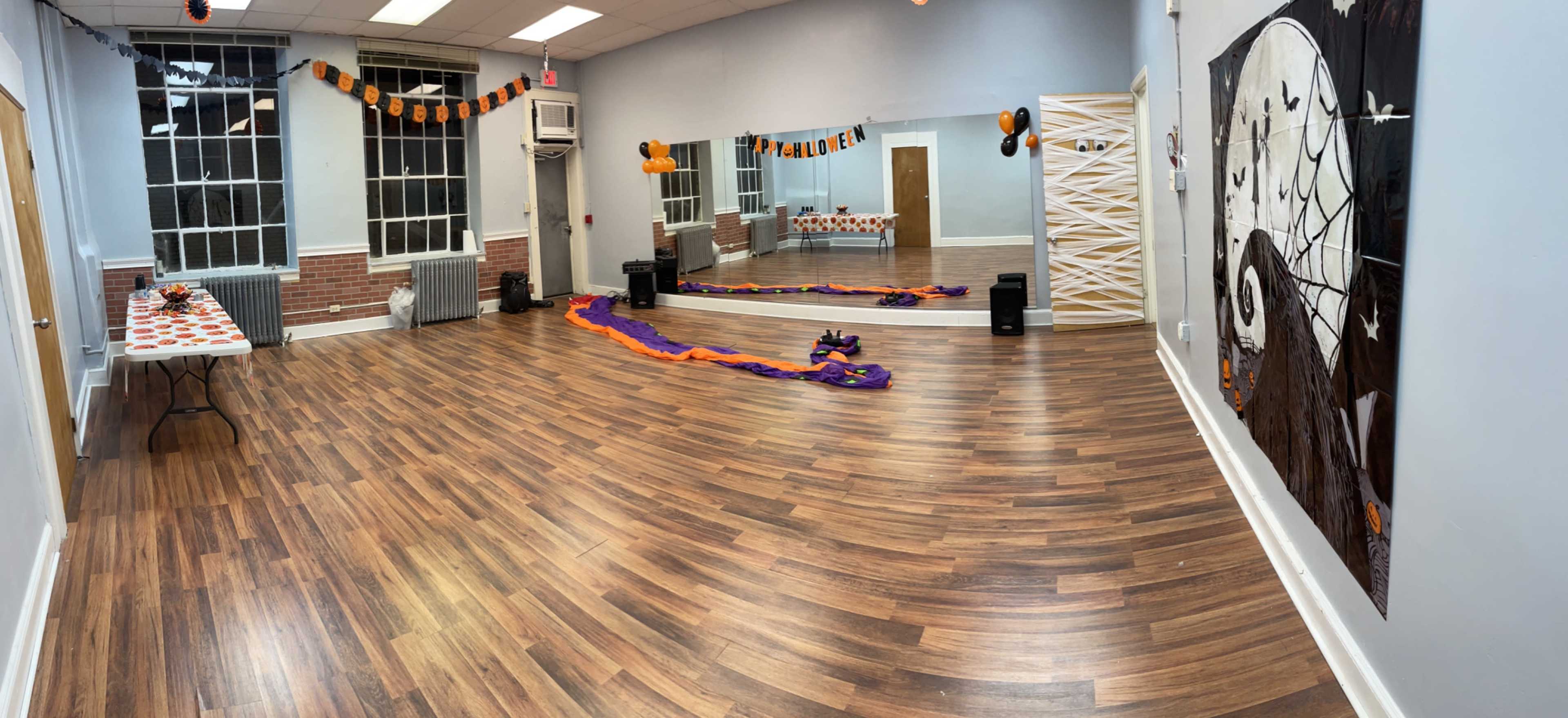 A spacious indoor room decorated for Halloween with orange and black decorations, a table with festive items, and purple streamers on the floor.