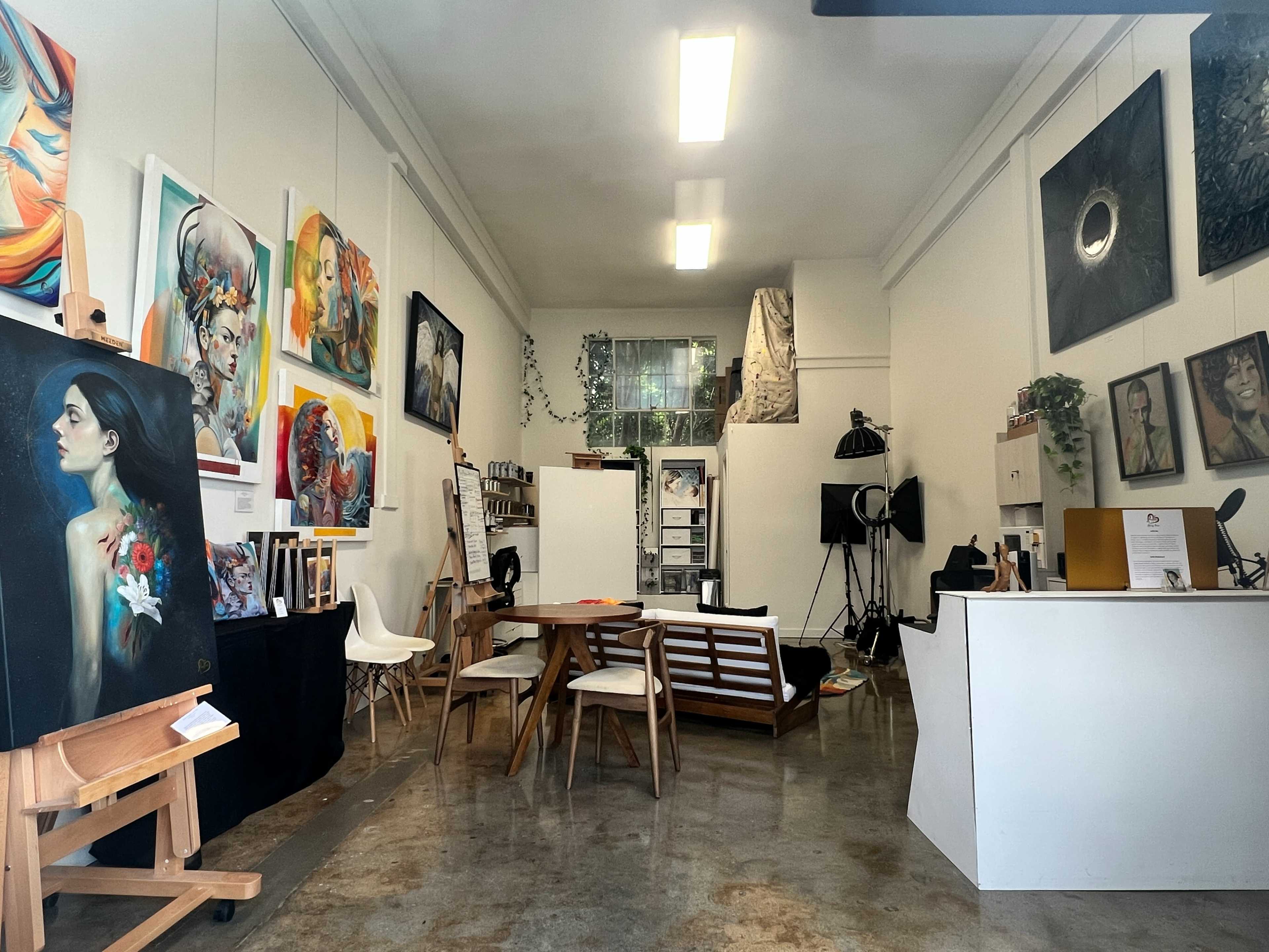 The image shows an art studio featuring several paintings on the walls, a wooden table with chairs in the center, and various art supplies and equipment scattered throughout the space.