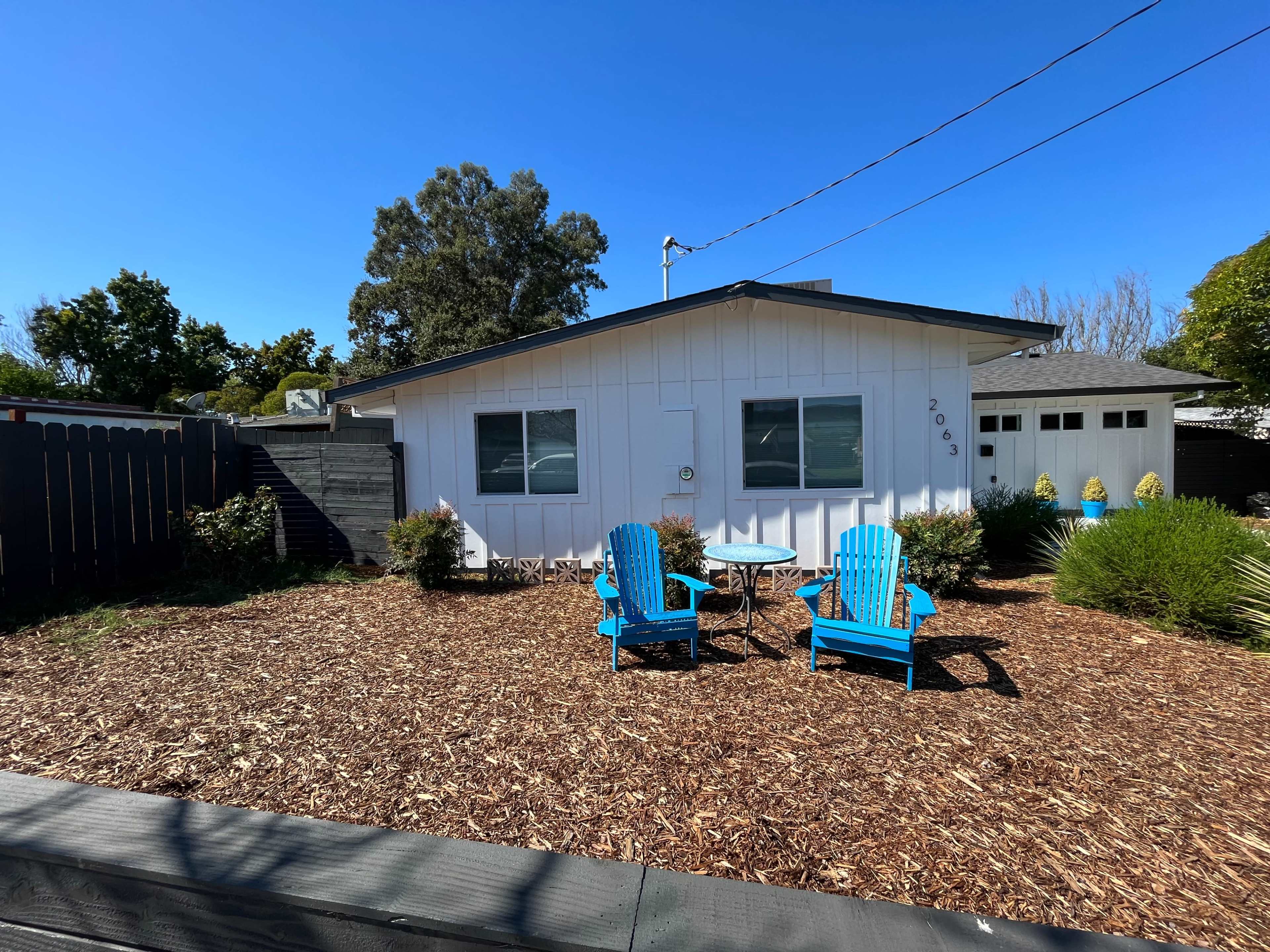 A small white house with a blue door and two bright blue chairs situated on a mulched front yard.