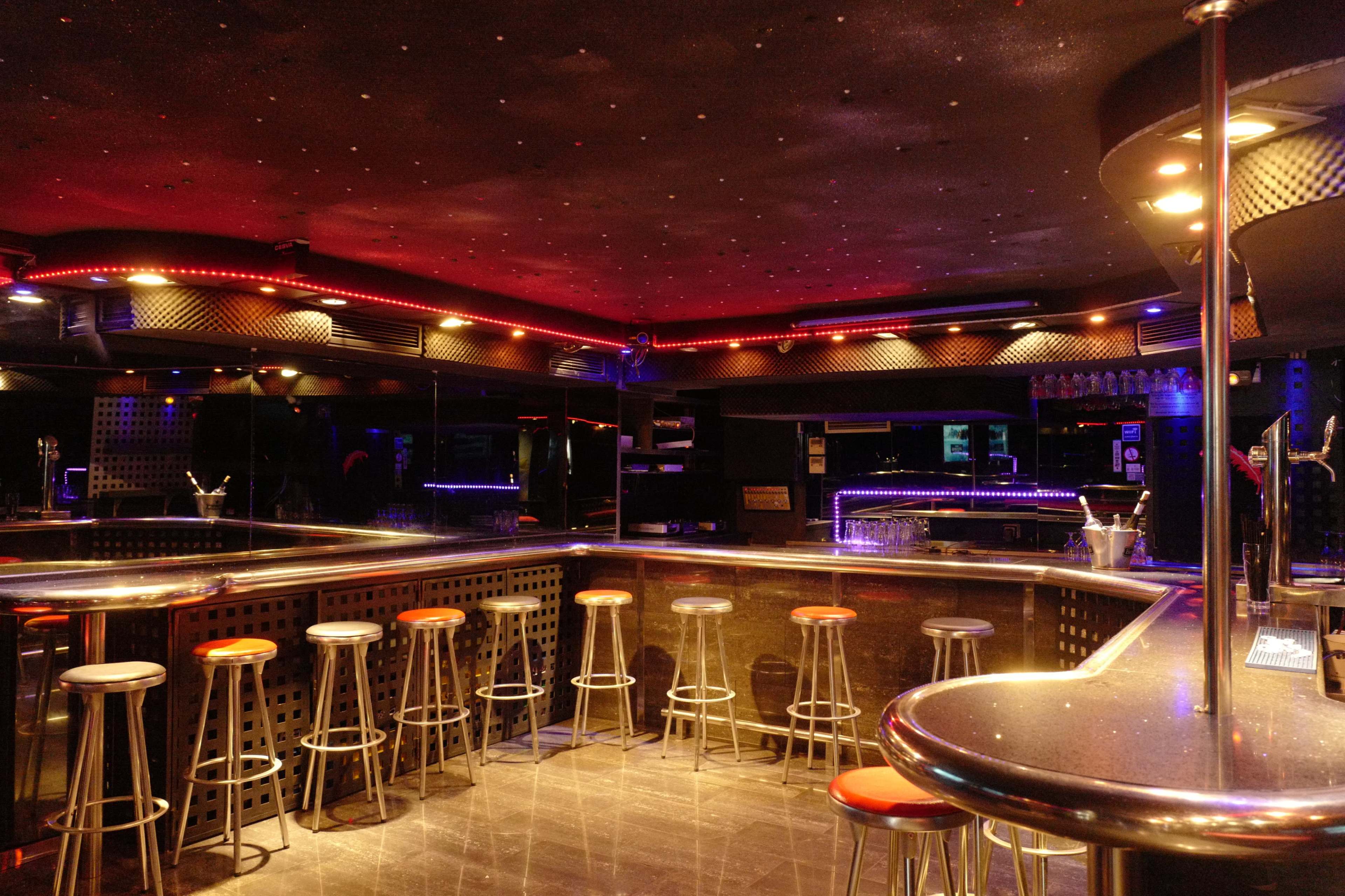 An empty bar area with high stools and a lit counter, featuring colorful lighting along the walls and ceiling.