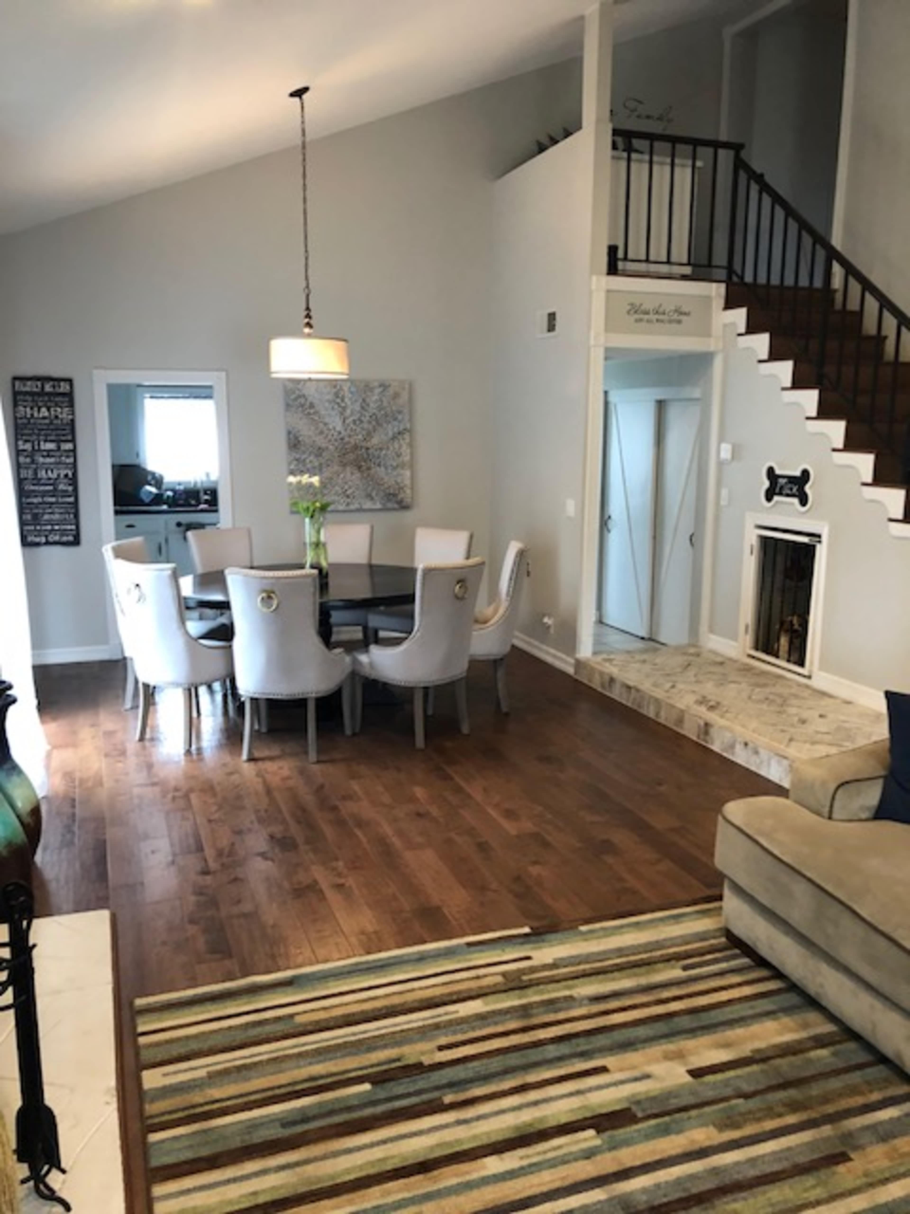 A dining area with a round table surrounded by upholstered chairs, adjacent to a staircase and a living area featuring a patterned rug and a beige sofa.