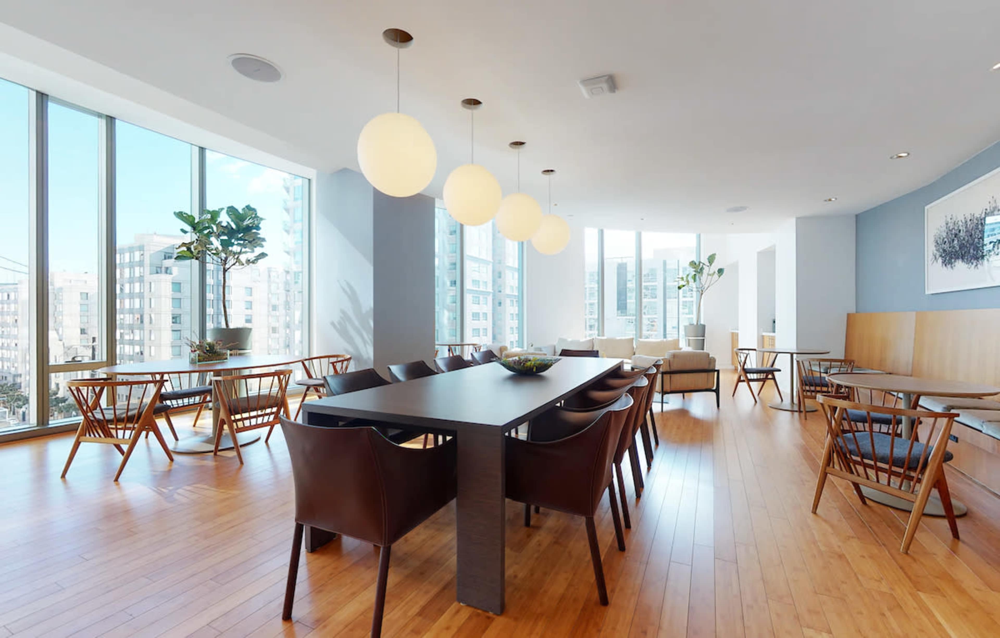 A modern dining area with large windows, featuring a long table surrounded by brown chairs and pendant lights.