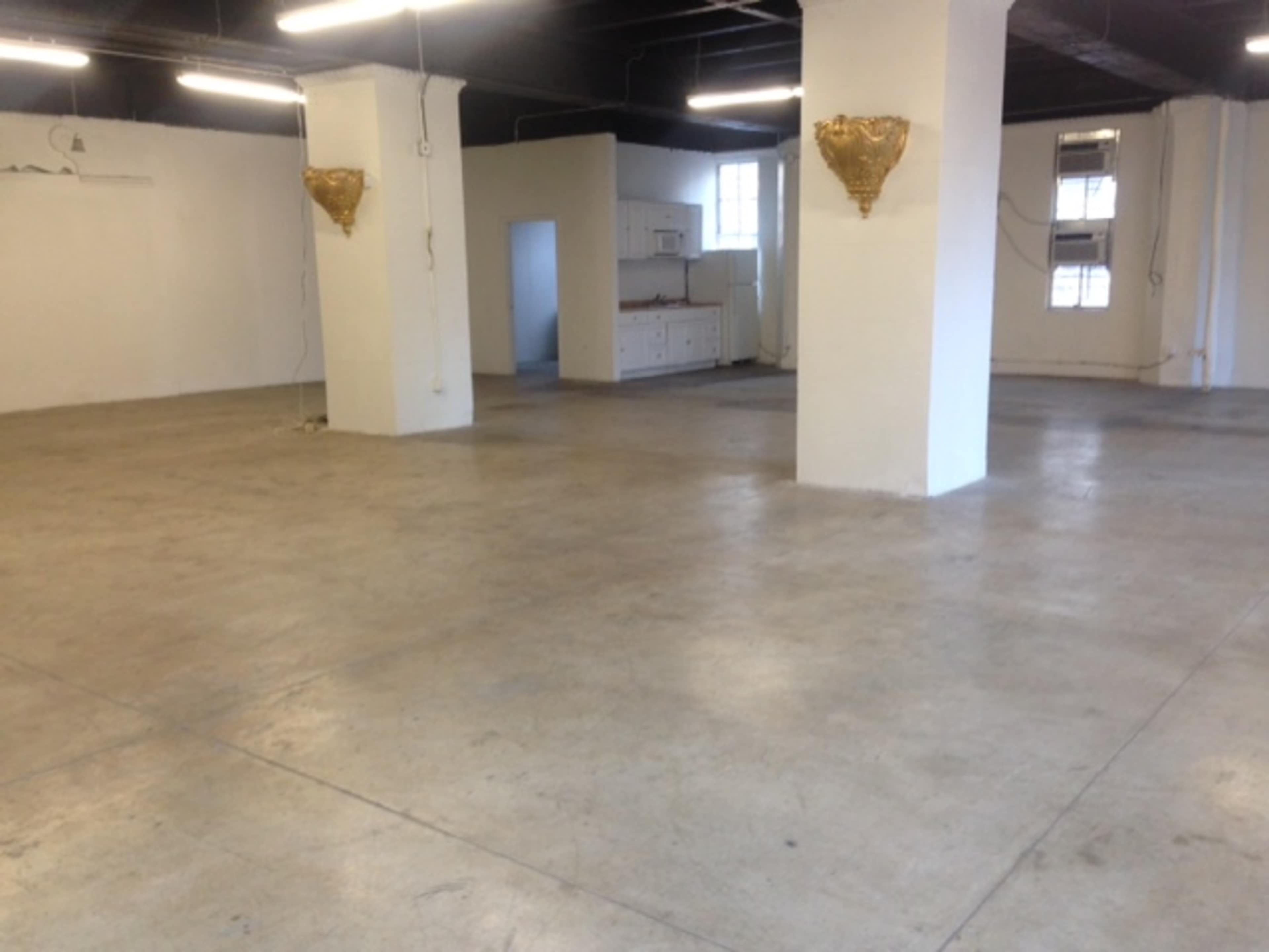A spacious, empty room with concrete floors and two ornate wall fixtures, leading to a kitchen area in the background.