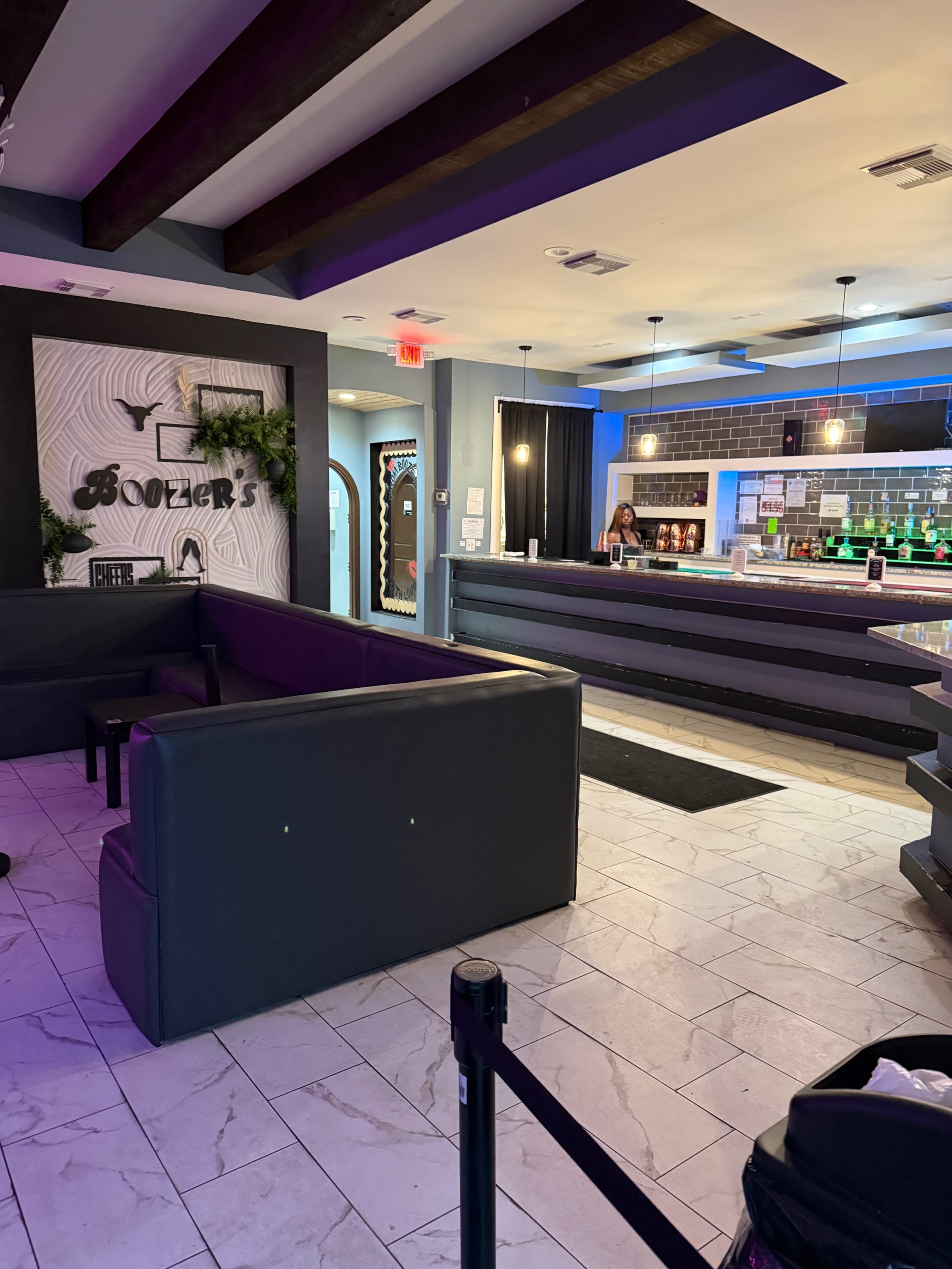 The image shows the interior of a bar with a spacious seating area, a long counter, and colorful lighting.