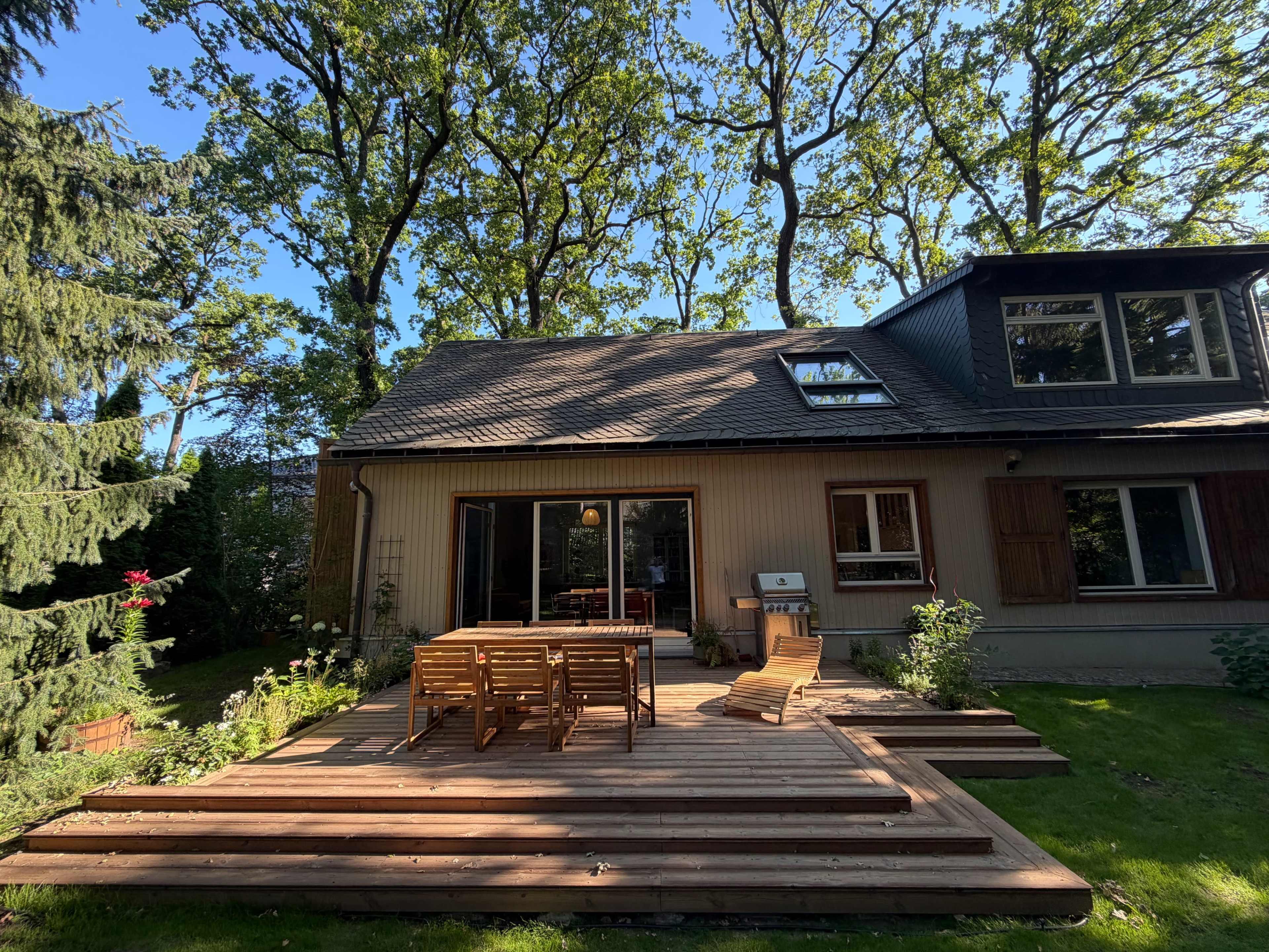 A wooden deck with seating overlooks a backyard surrounded by trees, adjacent to a two-story house.