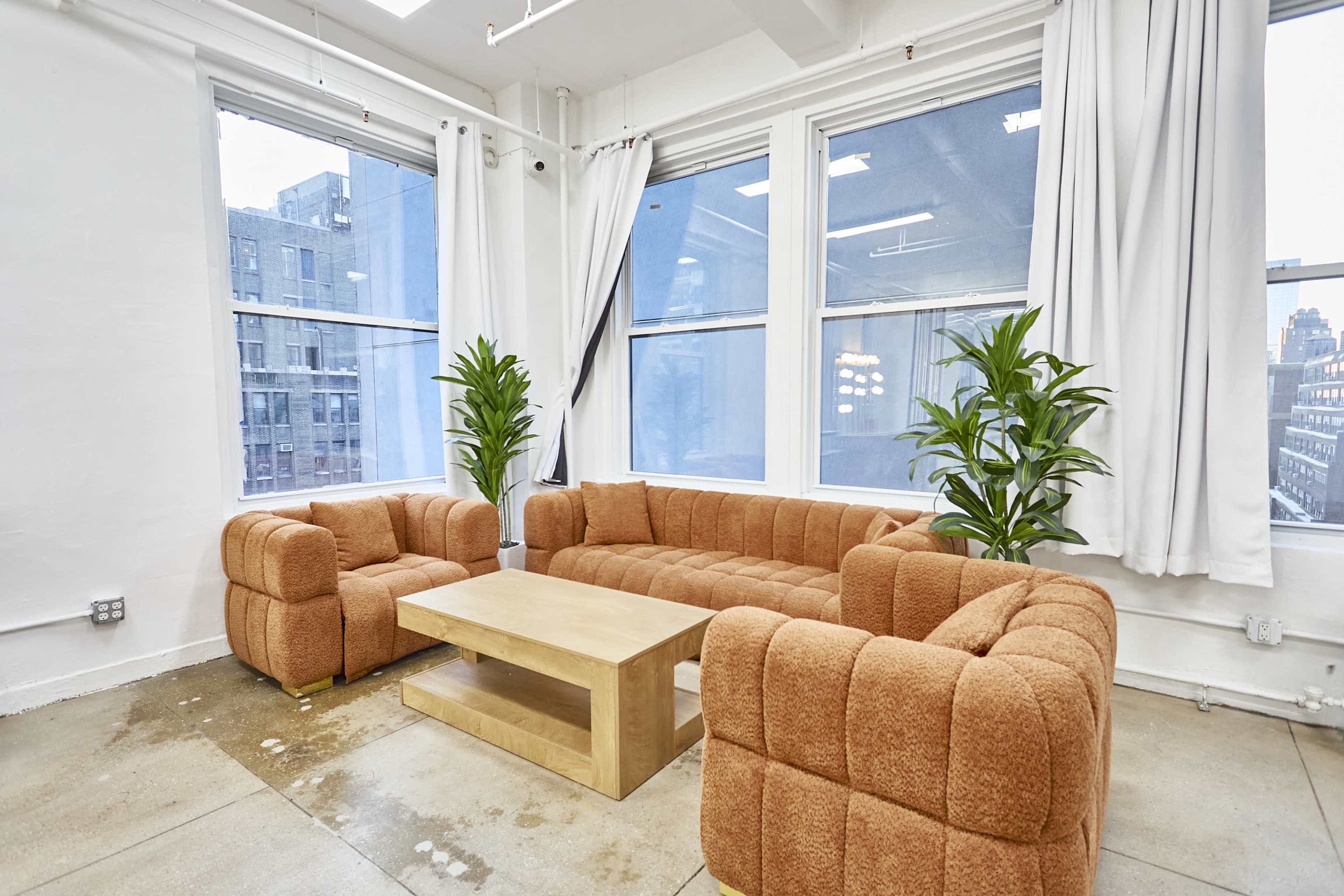 The image shows a cozy seating area with an orange upholstered sectional couch and a wooden coffee table, surrounded by large windows and indoor plants.