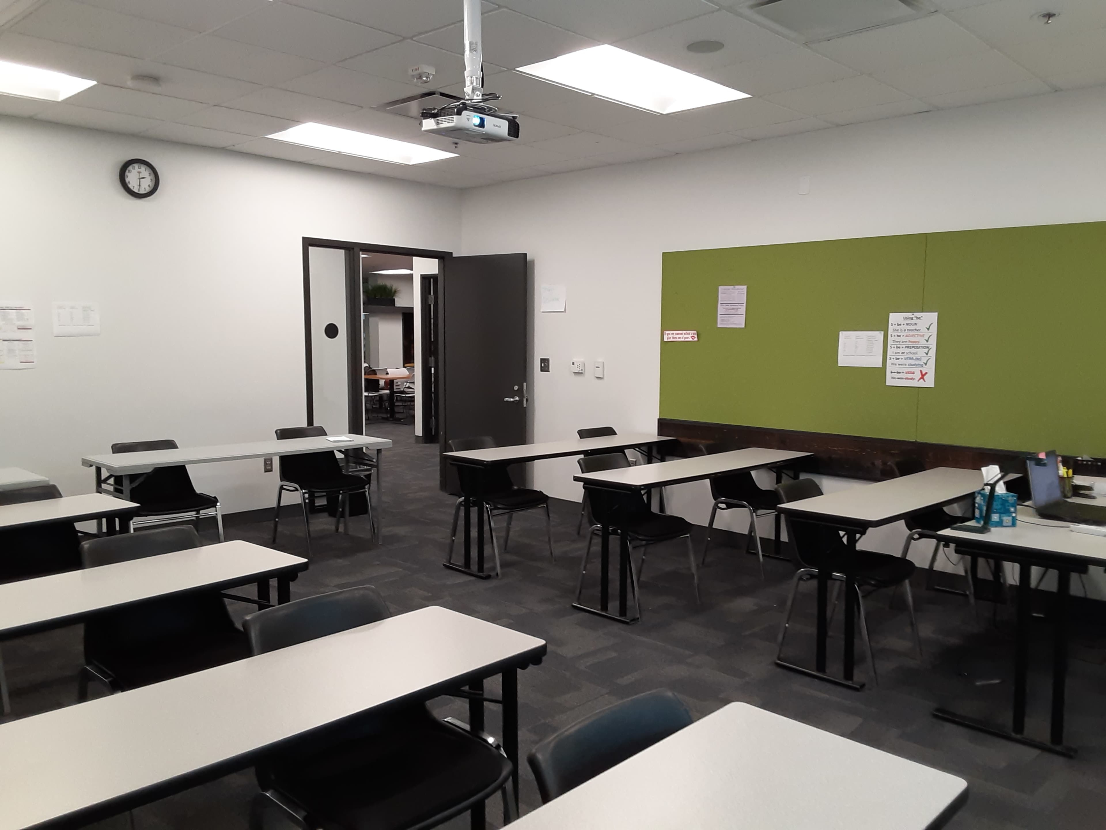 A classroom is set up with desks arranged in rows, a projector mounted on the ceiling, and a green bulletin board on the wall.