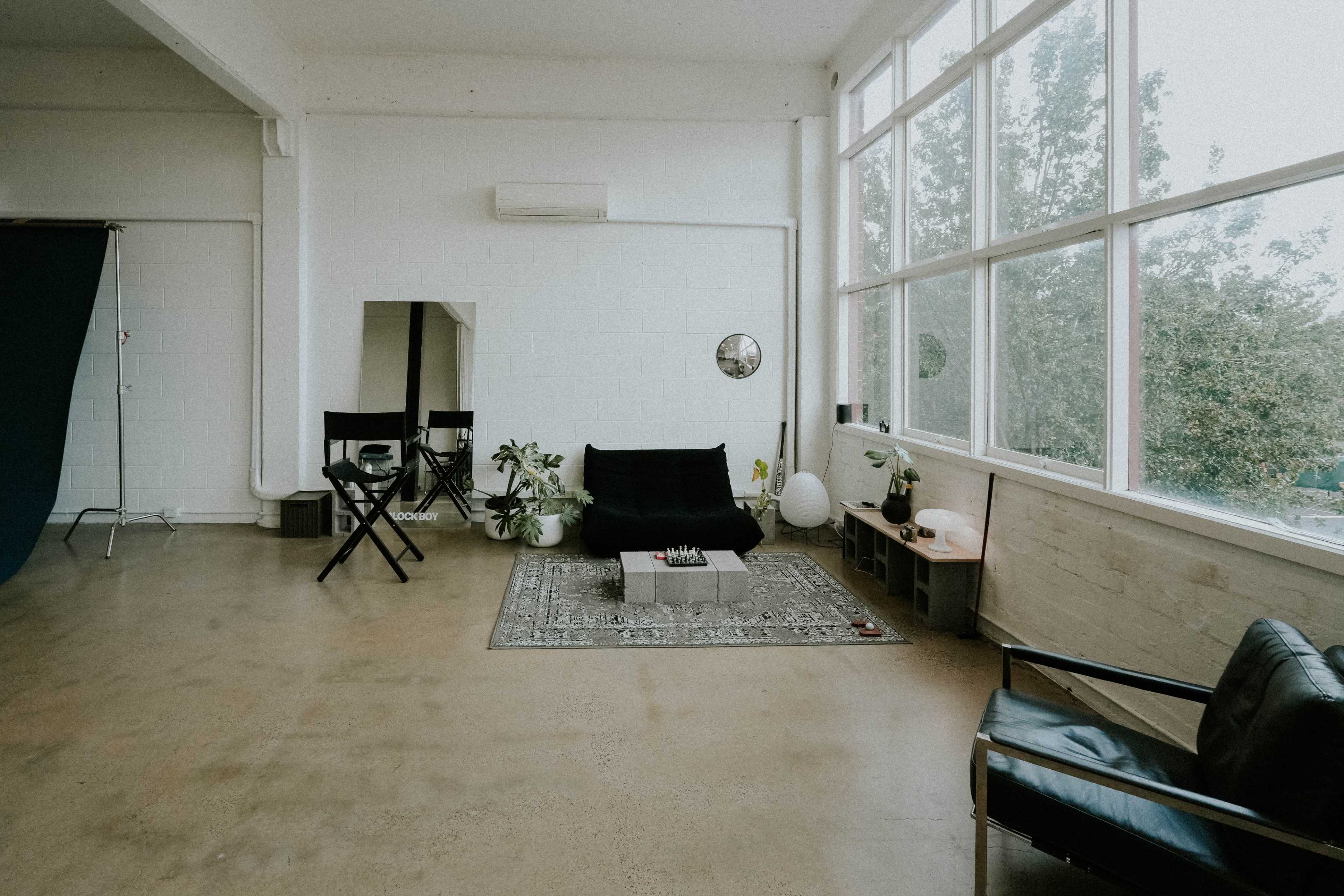 The image shows a spacious, simply furnished studio with large windows, a black sofa on a decorative rug, and various seating arrangements along with some potted plants.