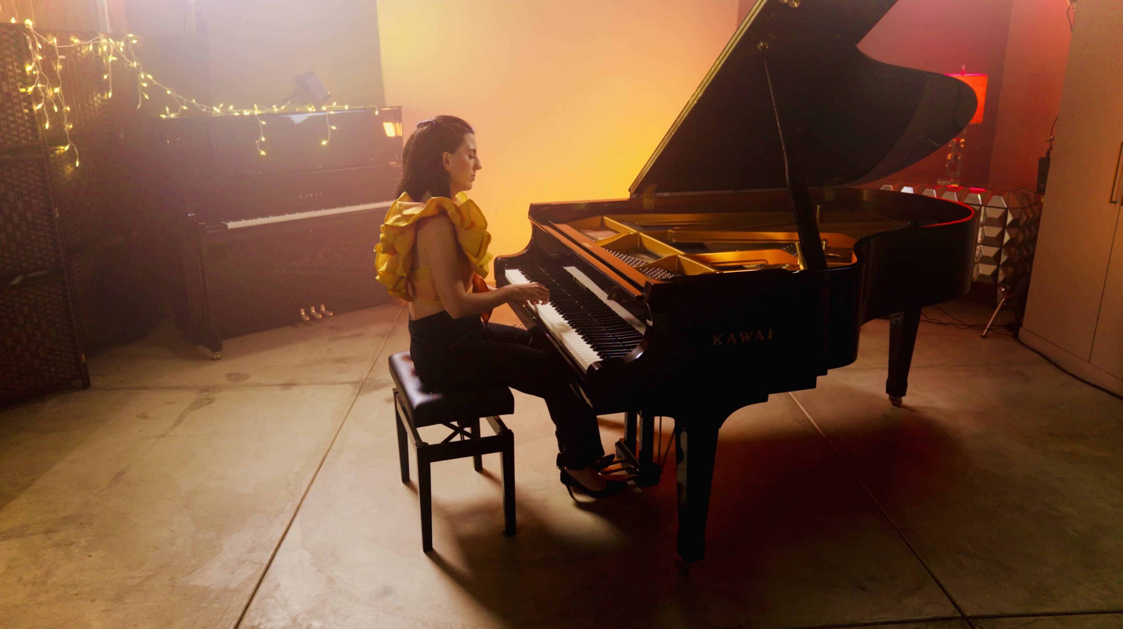 A woman in a ruffled yellow top plays a grand piano in a warmly lit room.