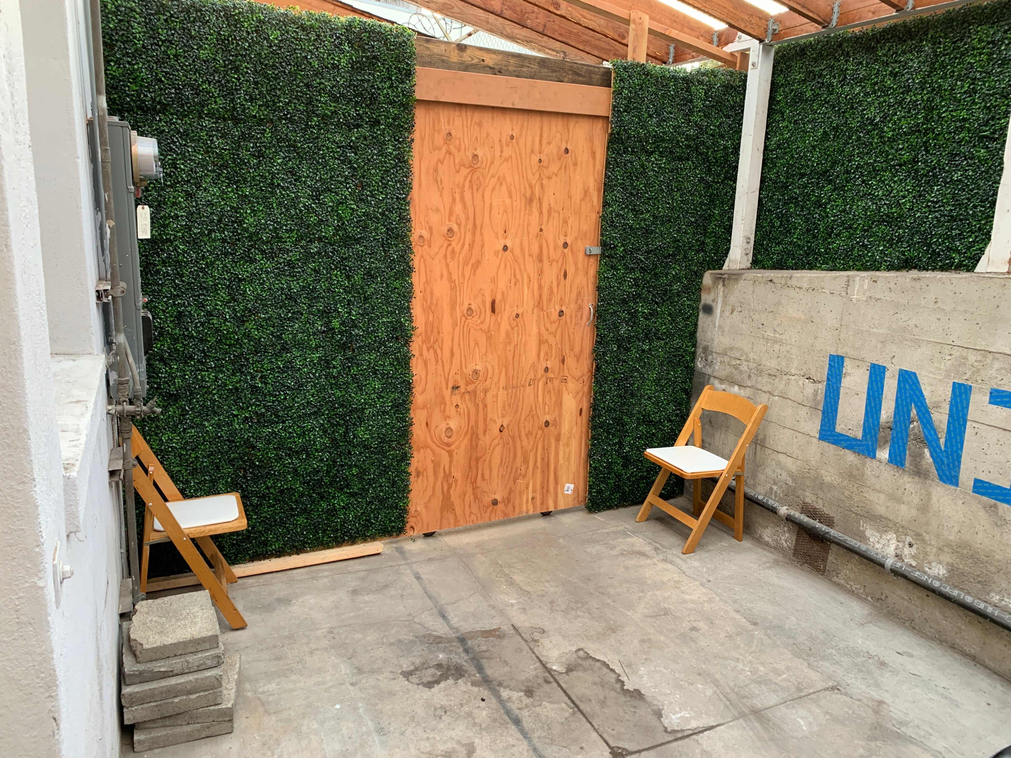A small, enclosed outdoor space with a wooden door, artificial greenery on the walls, and two wooden chairs positioned on a concrete floor.