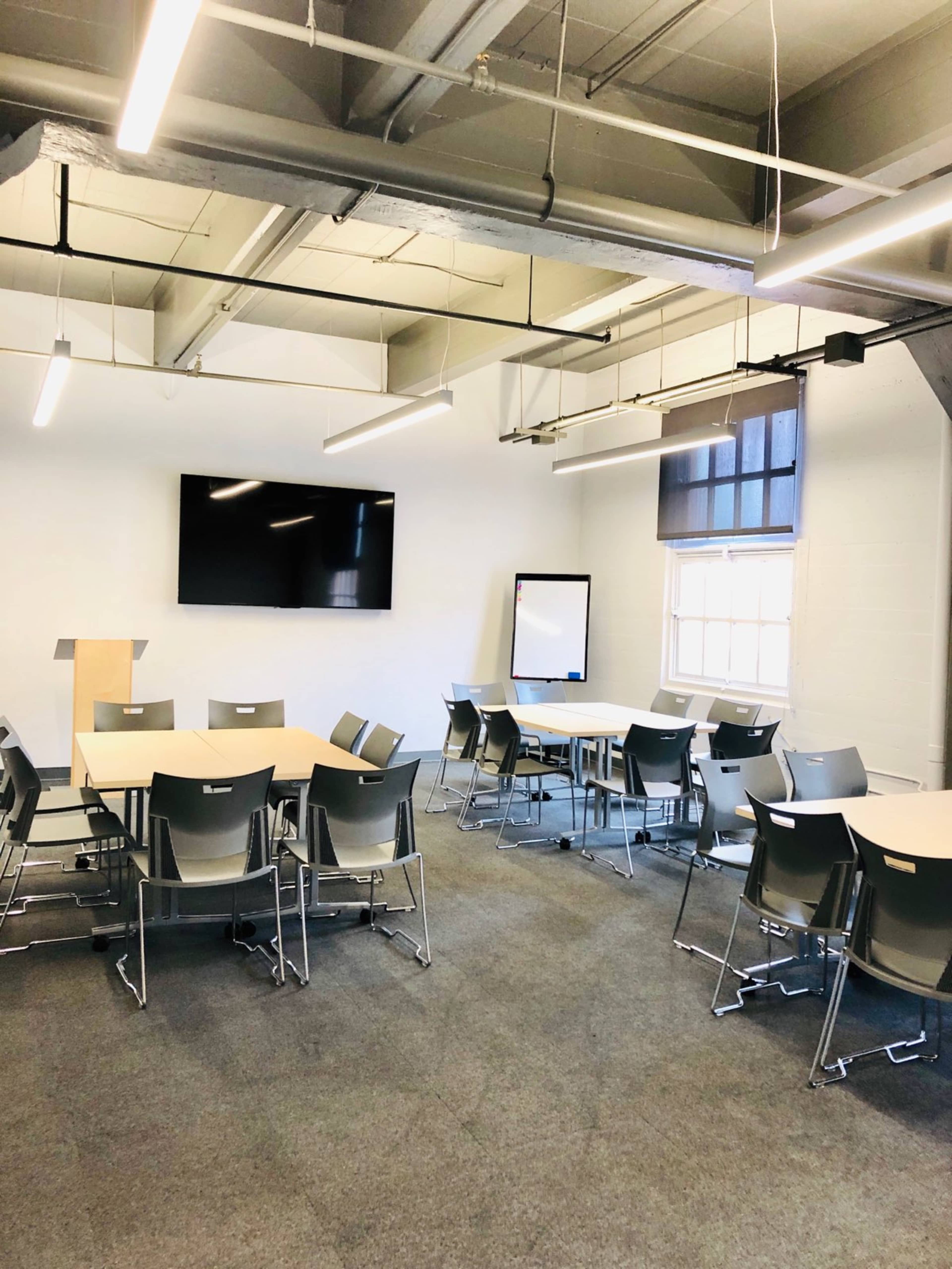 A spacious meeting room with multiple tables arranged in a U-shape, chairs, a large screen on the wall, and a whiteboard.