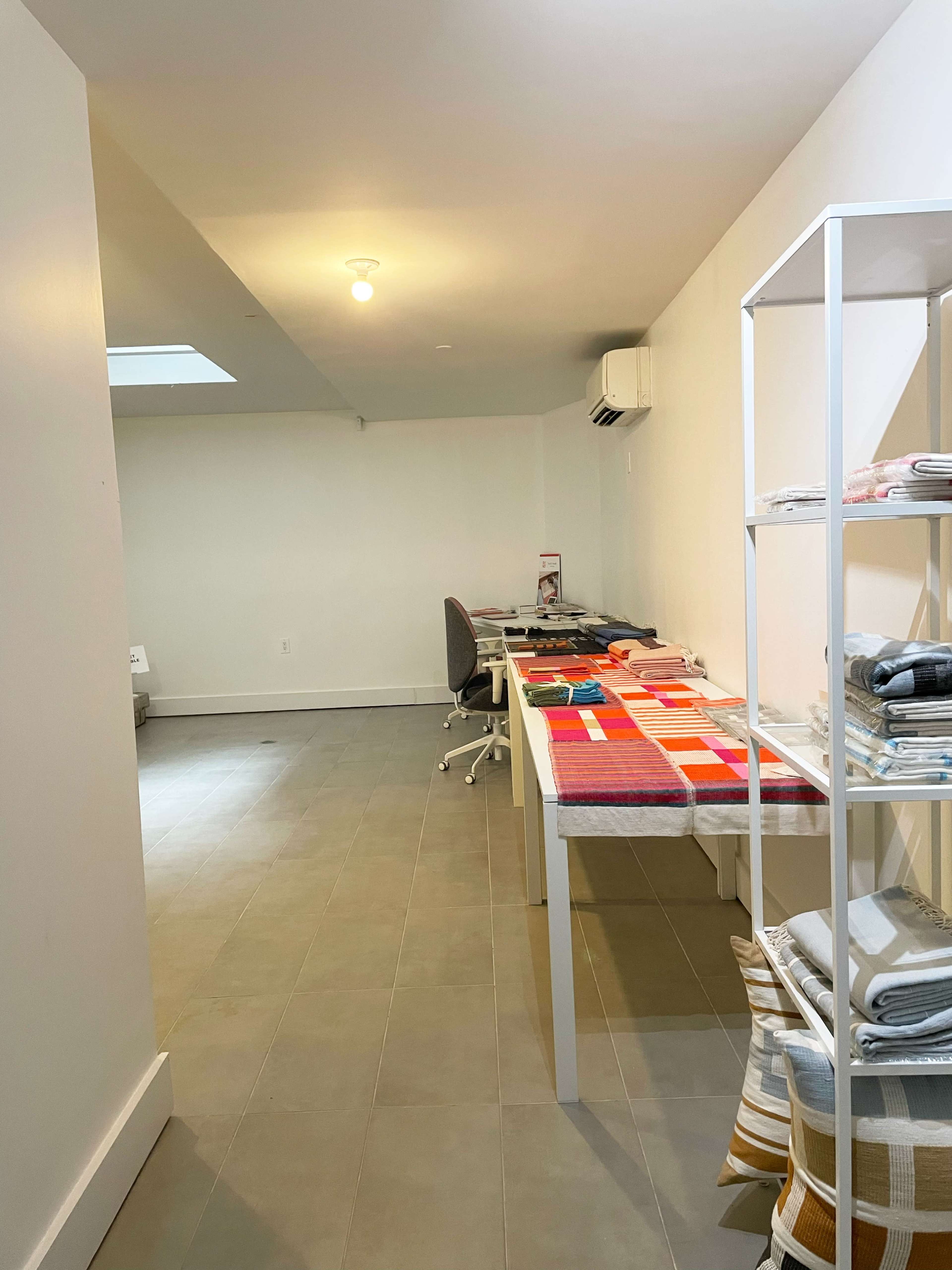 A spacious, well-lit room features neatly arranged textile products on tables and shelves, with a desk and chair visible in the background.