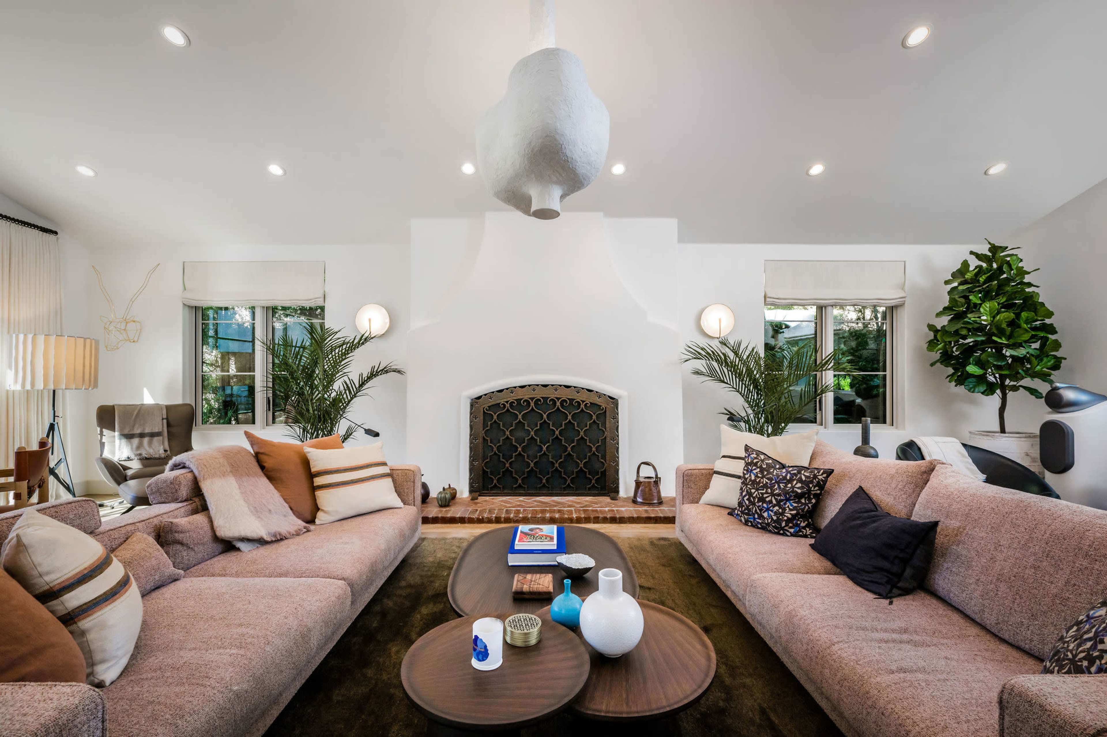 A spacious living room features two large sofas facing a central coffee table, with a decorative fireplace and plants in the background.