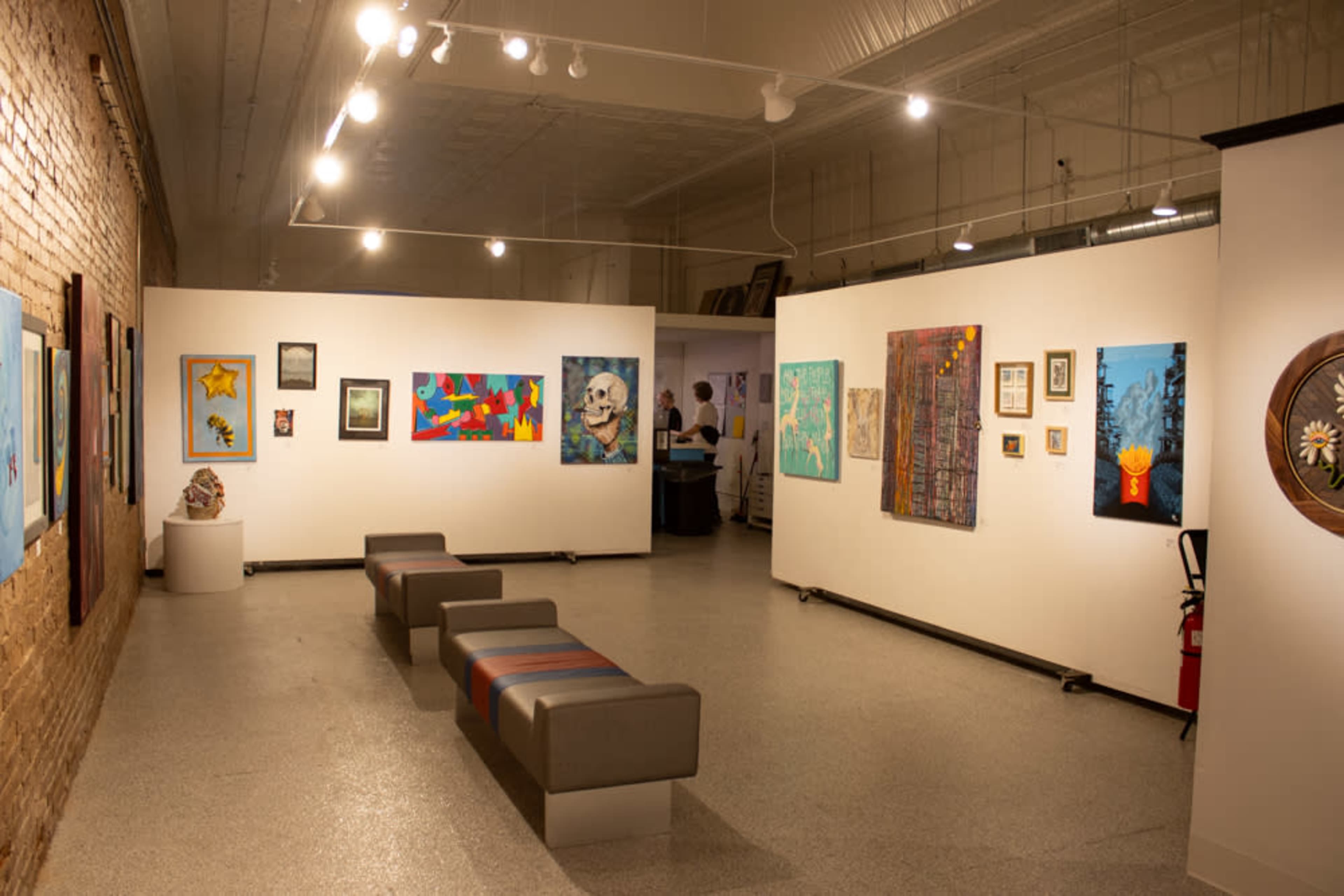 The image shows an art gallery with several colorful artworks displayed on white walls and a few gray benches arranged in the open space.